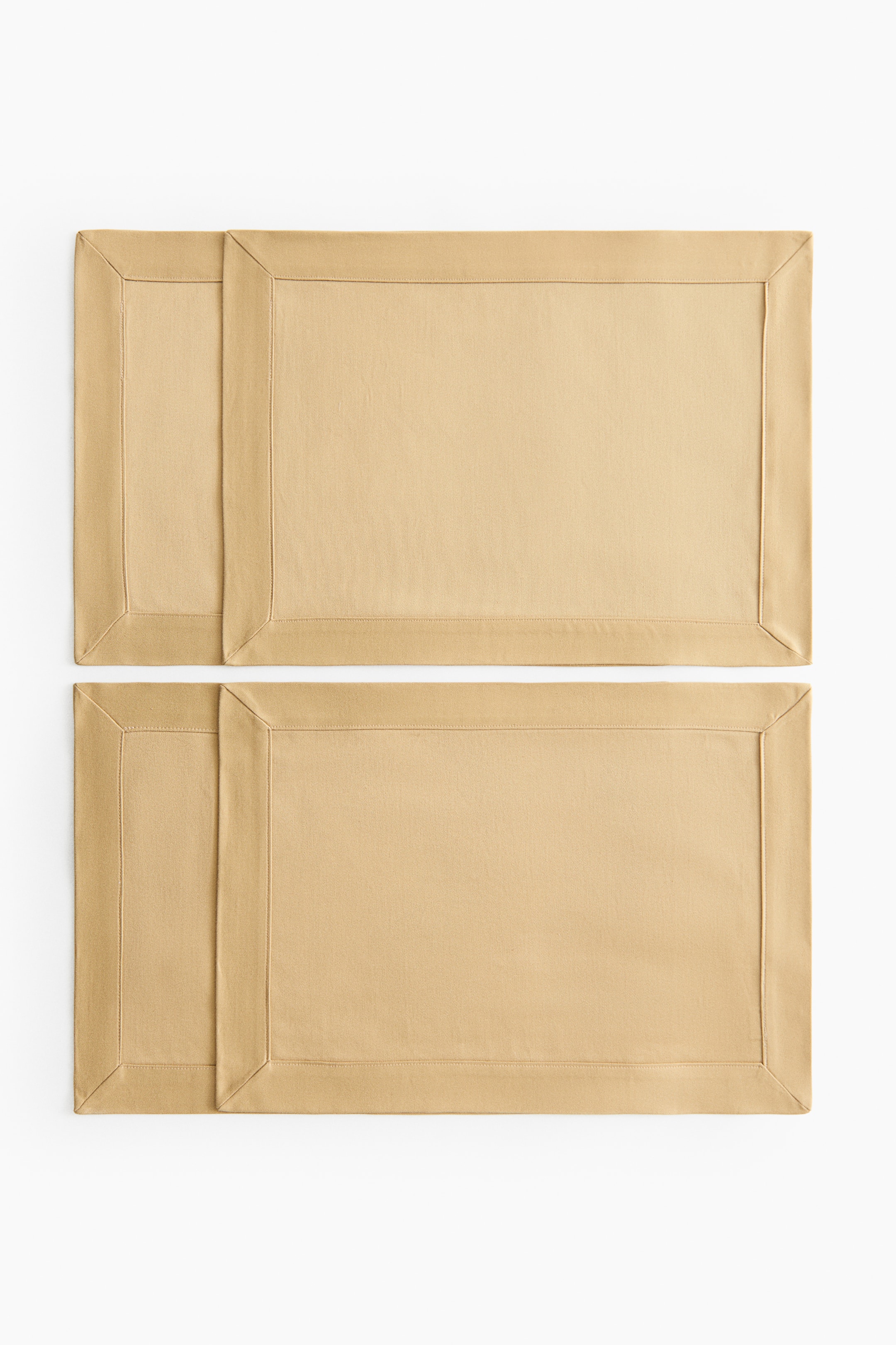 Ver imagen más grande: Cuatro manteles individuales rectangulares de color beige claro se muestran extendidos, cada uno con un amplio borde cosido y esquinas ingleteadas. Están dispuestos en dos filas horizontales, con el mantel individual de la izquierda en cada fila superponiéndose ligeramente al de la derecha.