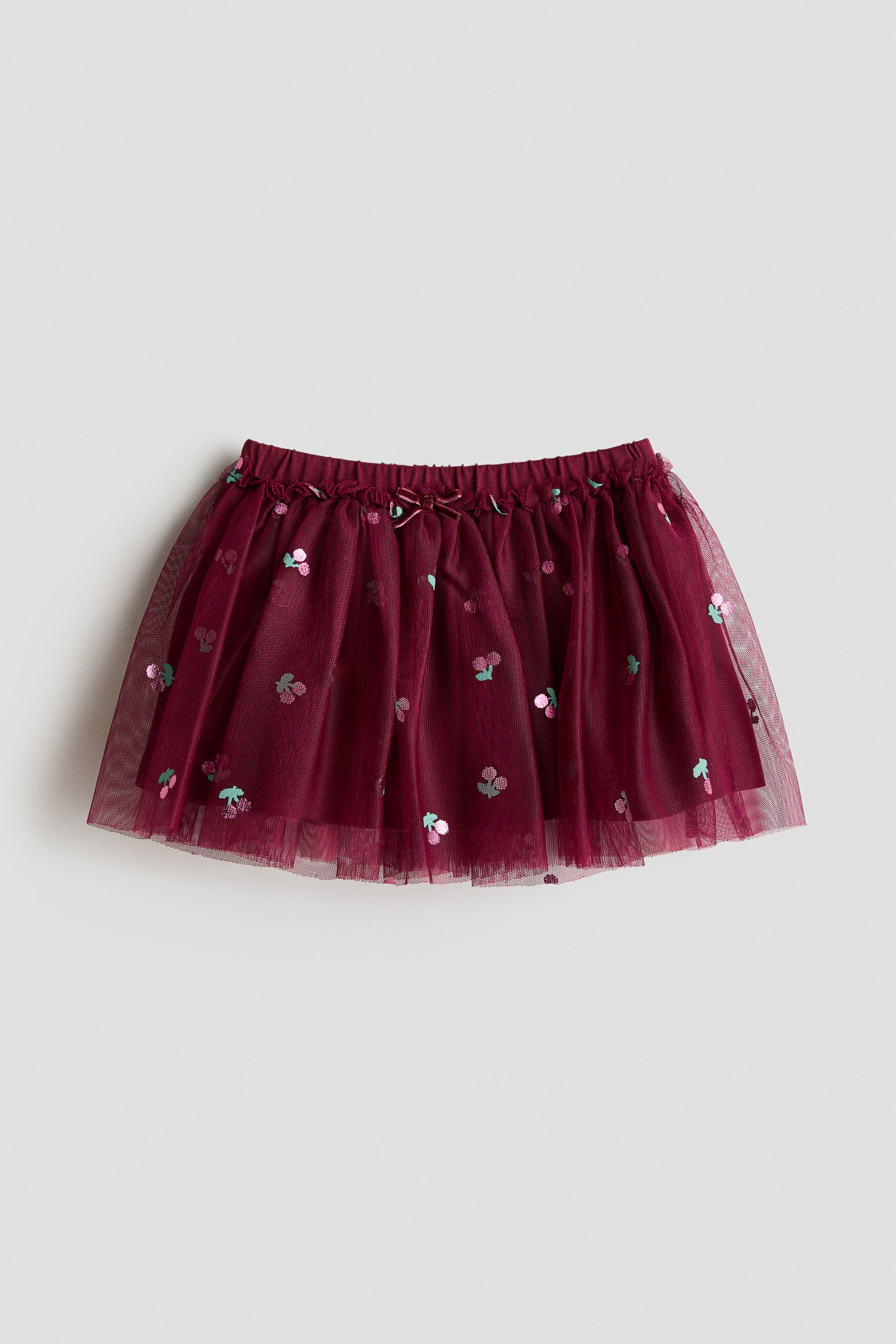 View larger image: A burgundy tulle skirt is showcasing a front view, featuring an elasticated waistband with a petite velvet bow at the centre. The skirt is embellished with scattered pink and green cherry motifs and boasts a flared hem.