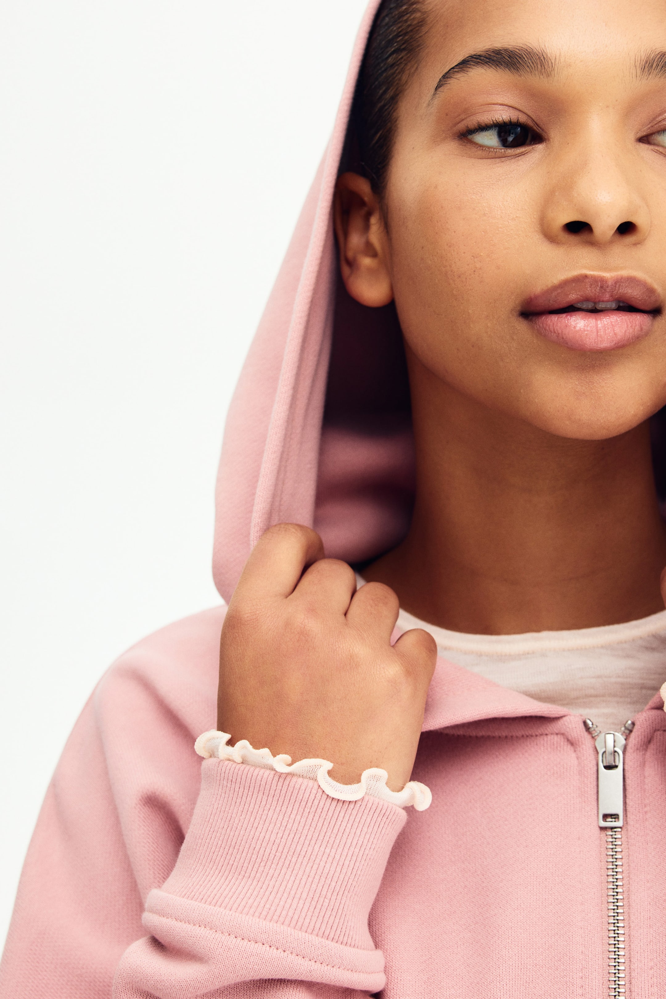 Grösseres Bild ansehen: Ein rosa Kapuzenpullover mit Reissverschluss, der über einen silberfarbenen Reissverschluss und gerippte Bündchen verfügt. Die Kapuze ist hochgezogen, wobei ein weisses Unterhemd am Halsausschnitt sichtbar ist und ein weisser Rüschenärmel unter dem Bündchen des Hoodies hervorschaut.