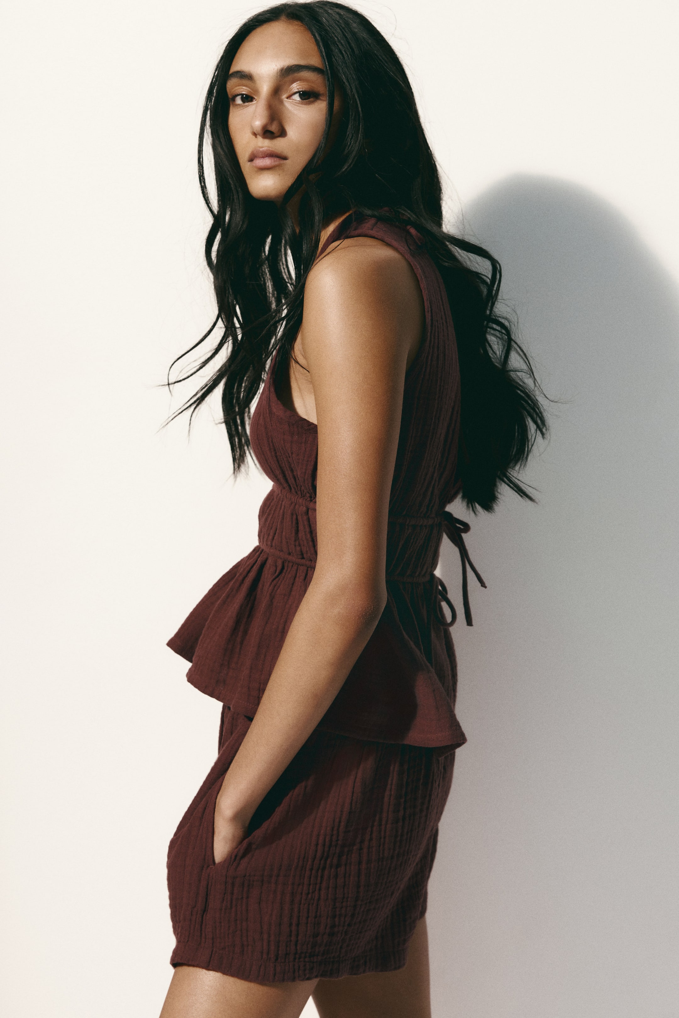 View larger image: A deep red crinkled gauze two-piece set is showcased from a right-facing angle. The sleeveless top includes a shirred waist that creates a ruffled peplum hem and ties at the back, while the matching shorts with pockets complete the ensemble.