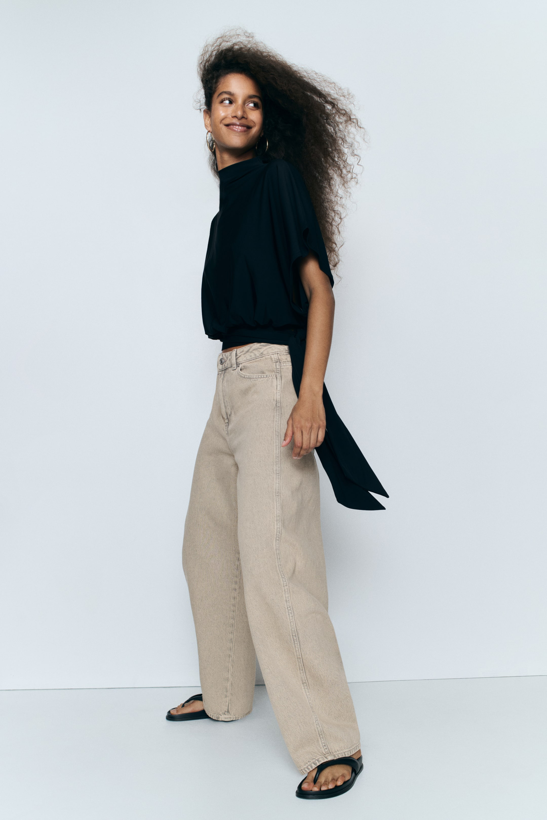 Grotere afbeelding bekijken: Lichtbeige denim jeans met hoge taille en een gebogen, volumineus model worden gedragen door het model. De jeans hebben vanaf de bovenbenen een losse, baggy pasvorm en zijn gestyled met een gedrapeerde zwarte top met hoge hals en zwarte platte sandalen.
