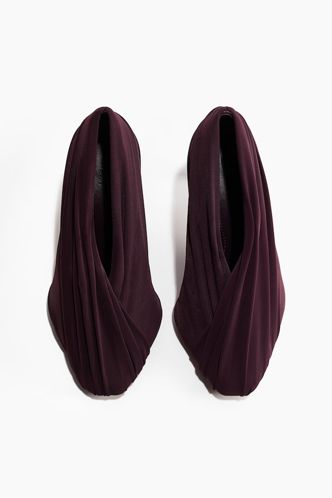 Zwei dunkelpflaumenfarbene Schuhe präsentieren sich aus der Frontansicht mit einem weichen, drapierten Stoffdesign. Der Stoff ist kunstvoll plissiert und um die Form des Schuhs gelegt, wodurch ein skulpturaler Effekt rund um den Schuhausschnitt und die abgerundete Schuhspitze entsteht. - 1