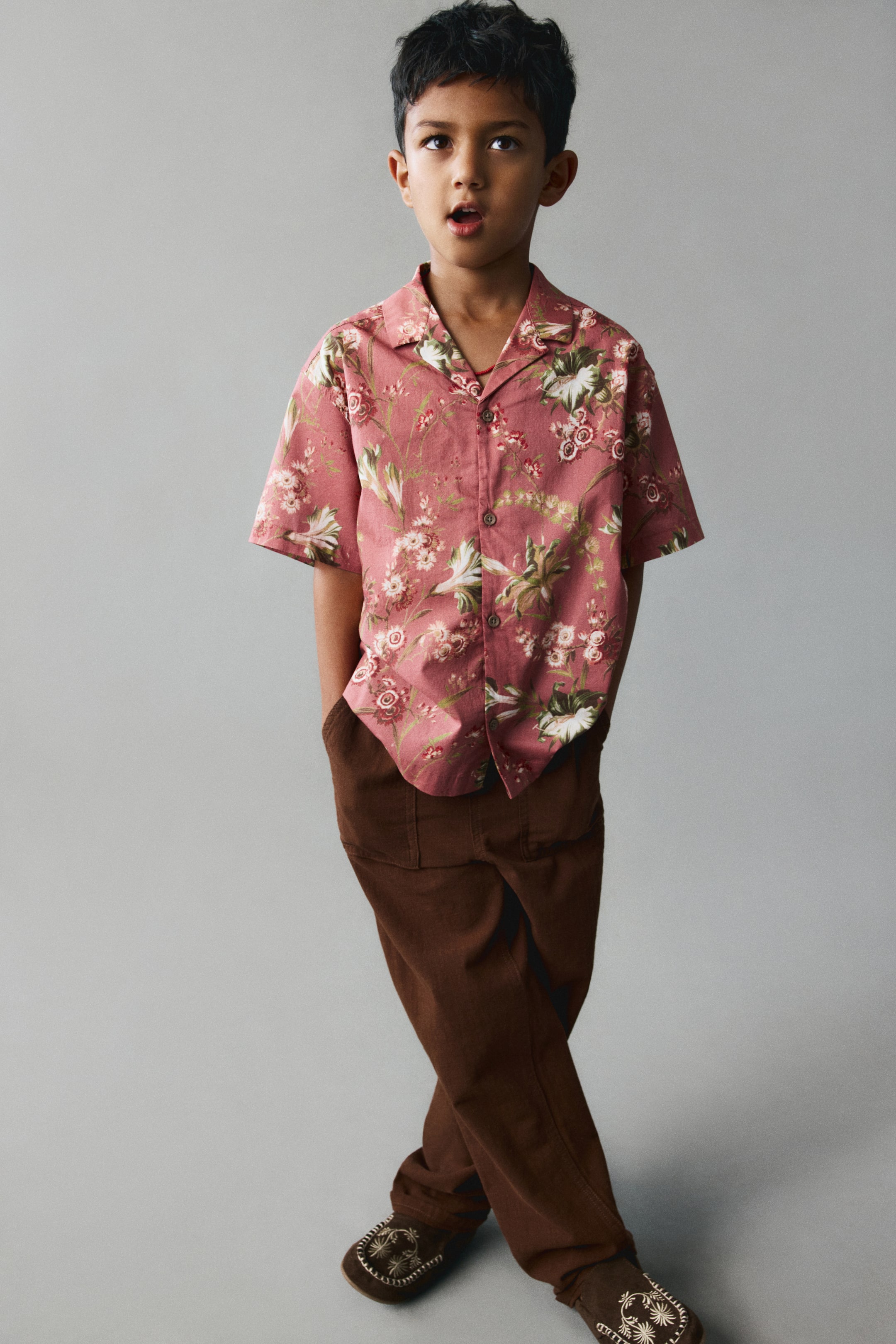 View larger image: A short-sleeve shirt in dusty pink cotton weave with an all-over floral print is worn facing front. The shirt features a resort collar, a button-down front, and gently dropped shoulders, draping loosely over brown trousers.