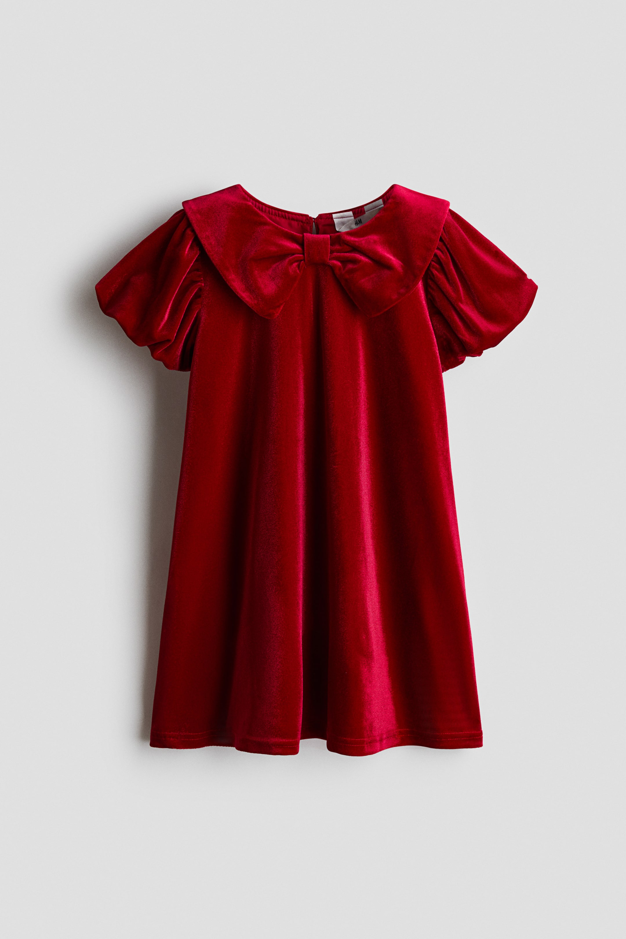 View larger image: A red velvet dress is shown from the front, featuring a wide collar adorned with a large matching bow at the centre neckline. The dress features short puffed sleeves and a flared A-line silhouette with soft draping.