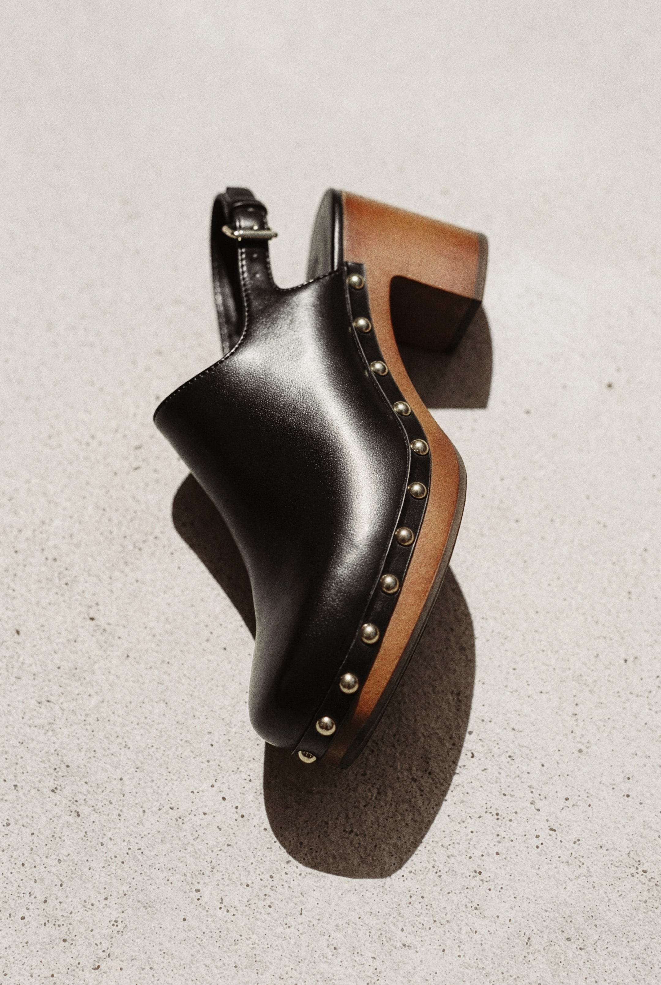 View larger image: A black leather clog is showcased in its side profile, highlighting a sleek black leather upper with a prominent black leather slingback strap and buckle. Gold-toned studs embellish the junction between the upper and the light brown wooden platform sole, which elevates into a coordinating block heel.