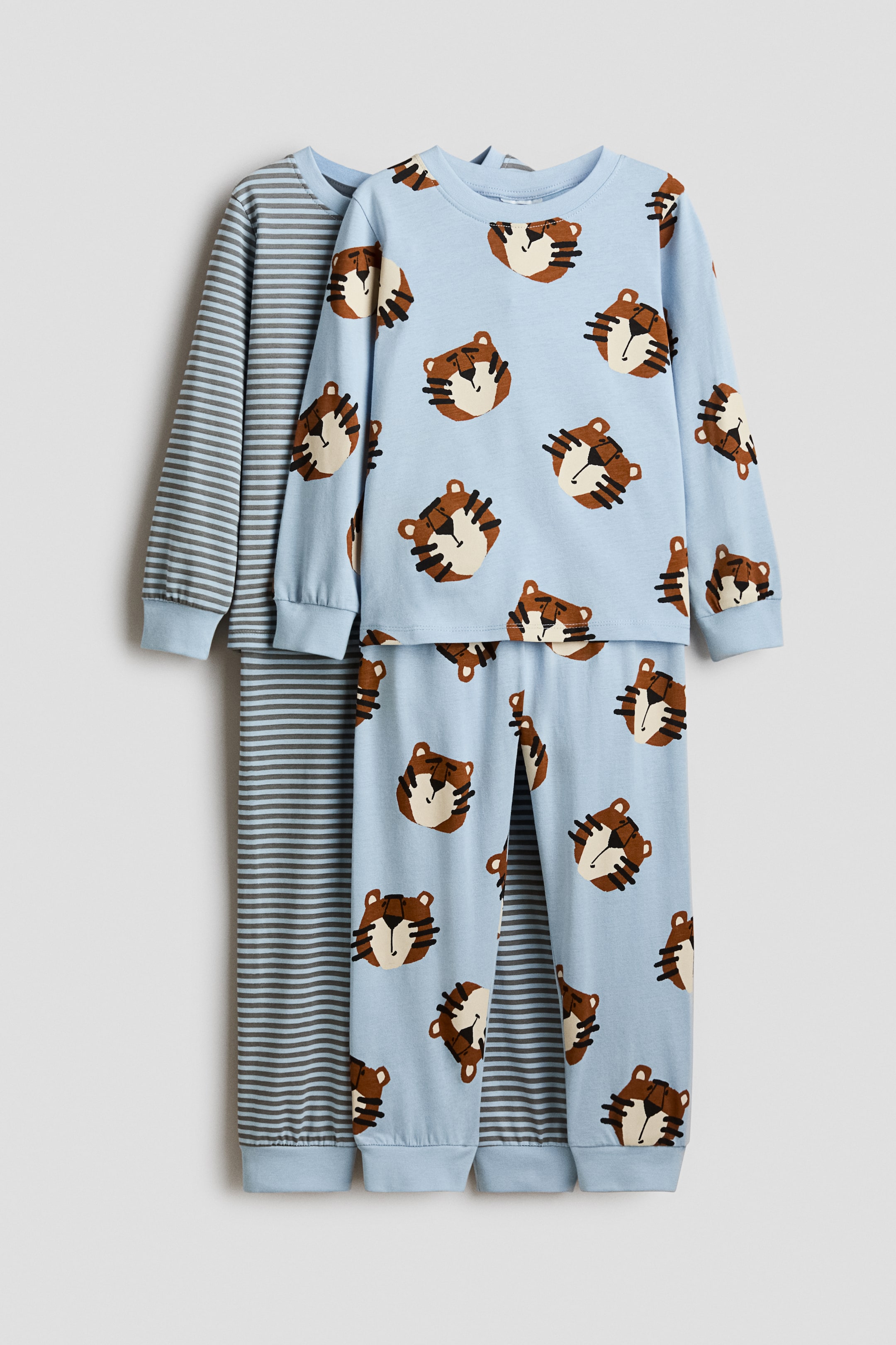 View larger image: Two sets of pyjamas in soft, printed cotton jersey are shown from the front, both featuring long-sleeved tops with round necklines trimmed in rib, and ribbed cuffs, and bottoms with elasticated waists and ribbed hems. The top set is light blue with an all-over print of brown tiger faces, while the partially visible set underneath features light blue and grey stripes.