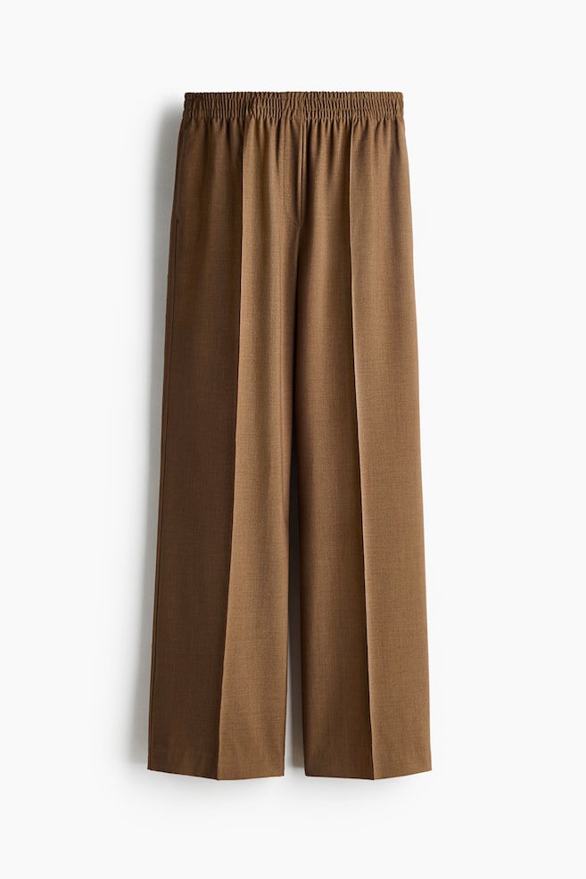 Pantalon à coupe ample en tissu tissé brun, présentant une taille haute avec élastique smocké, une fausse braguette à l'avant, des poches latérales discrètes et des jambes larges ornées de plis cousus sur le devant. - 1