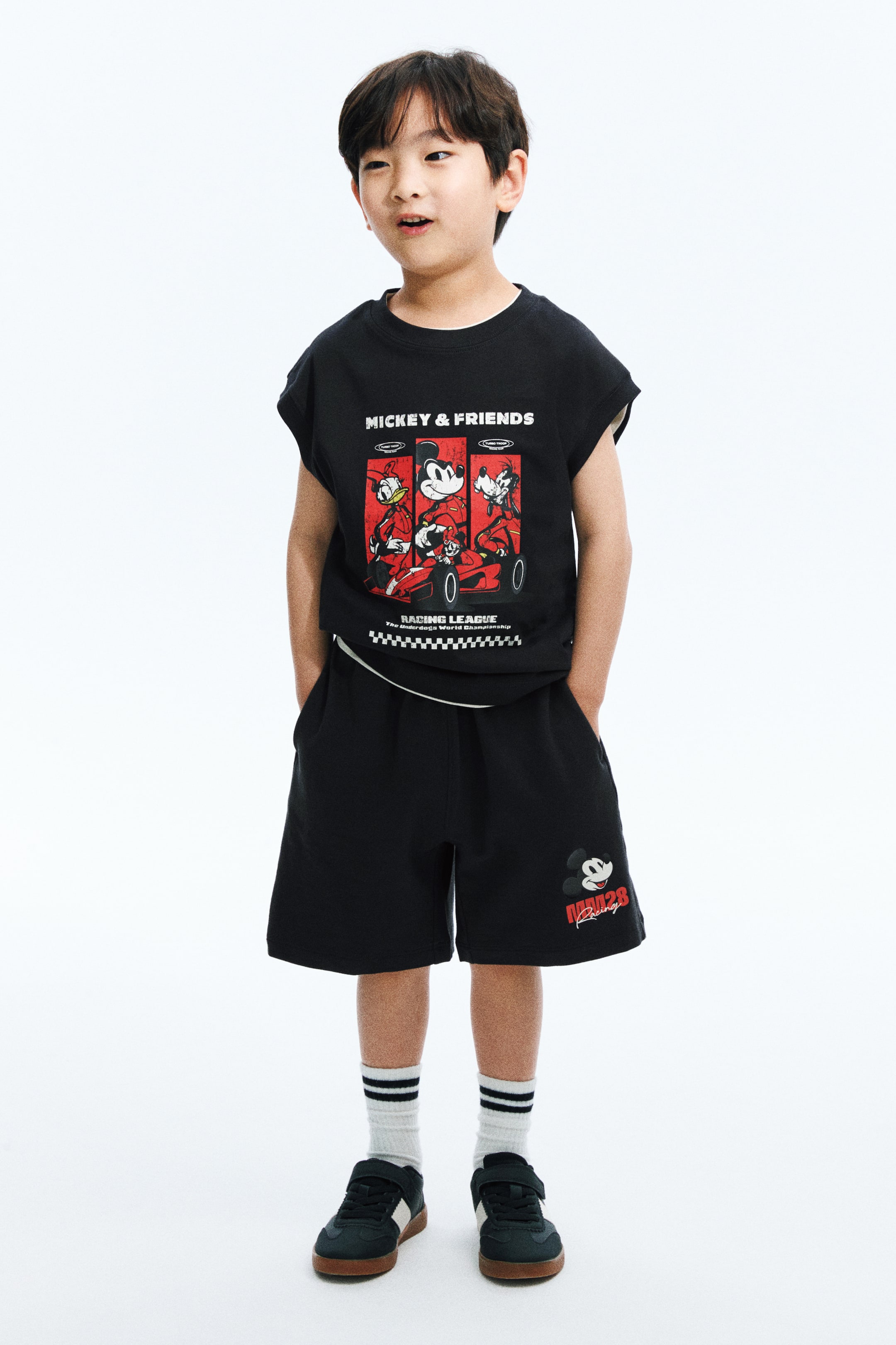 View larger image: A black two-piece set, facing forward, features a sleeveless crew-neck t-shirt and knee-length shorts, both with a relaxed fit. The t-shirt showcases a "Mickey & Friends" racing graphic with Donald, Mickey, and Goofy in red race cars, along with "Racing League" text and a checkered flag border. The shorts display side pockets and a small Mickey Mouse head with "RACING" text on the left leg.