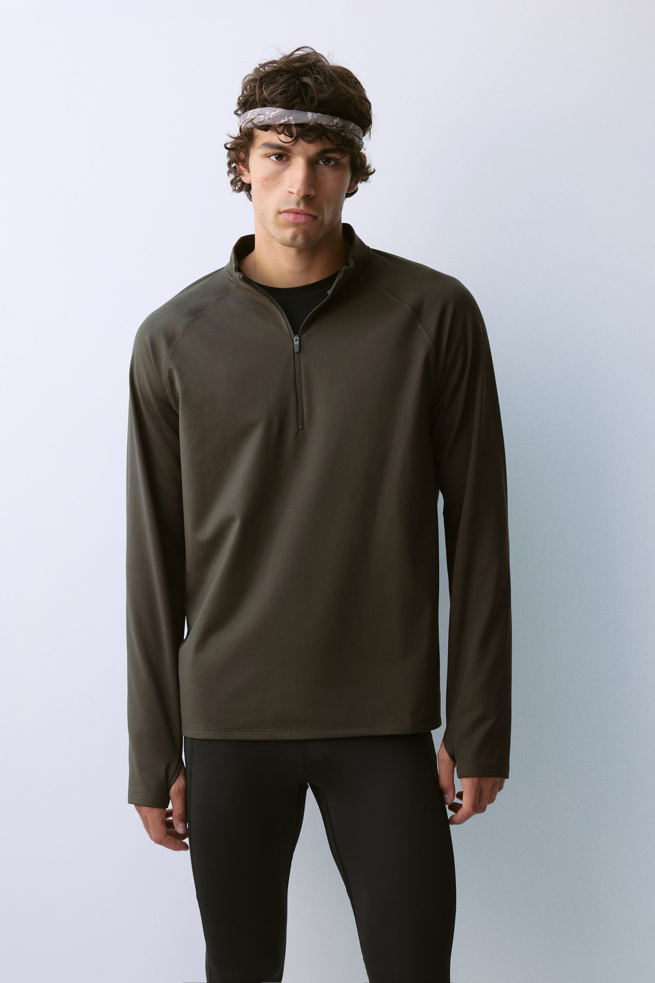 View larger image: A regular-fit half-zip activewear top in dark olive green is worn by the model, featuring long raglan sleeves, a high collar, and a partially open zipper. The top is layered over a black crew-neck T-shirt and paired with black athletic tights.