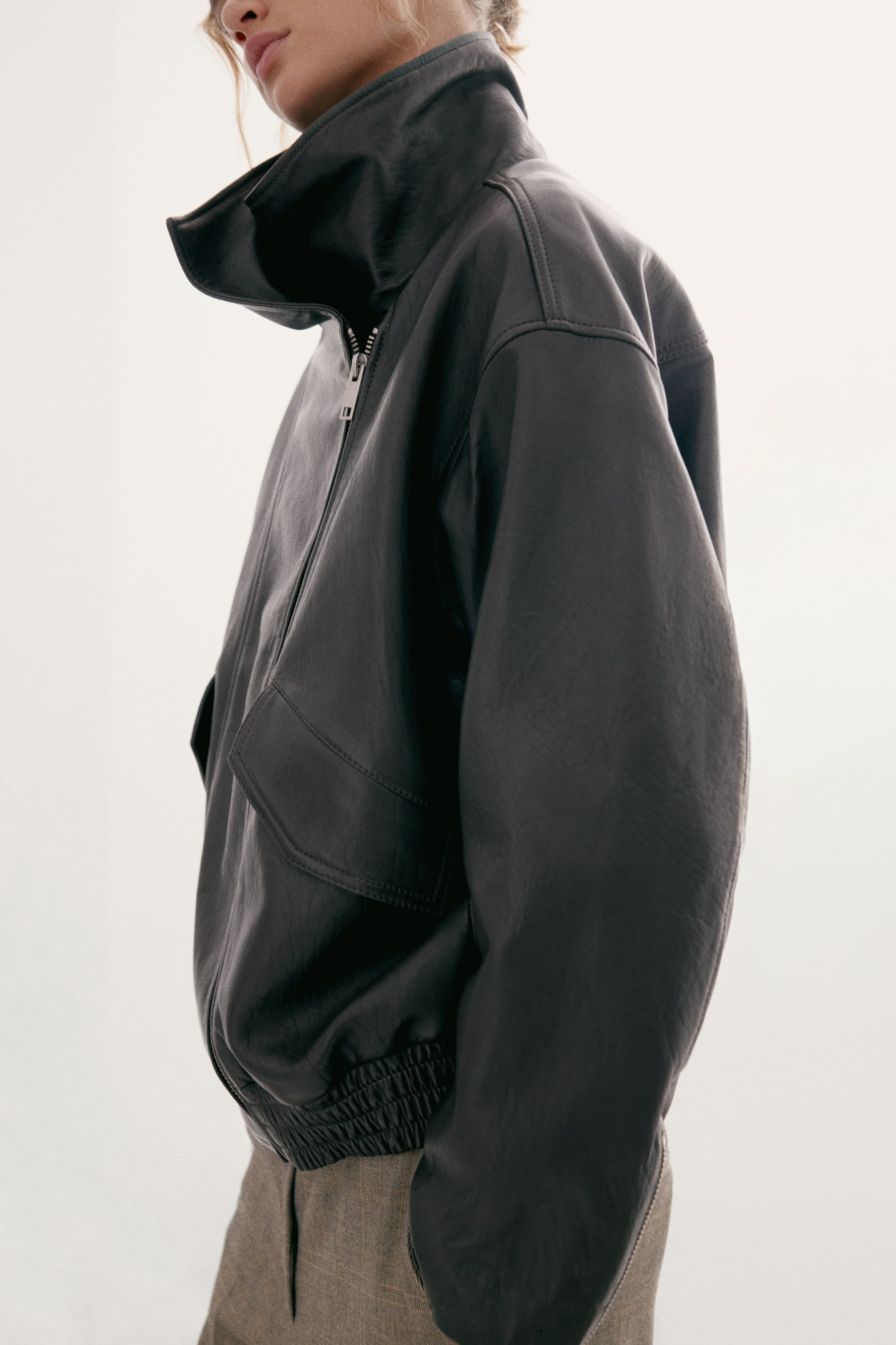 Ver imagen más grande: La modelo lleva una chaqueta de cuero oscuro de corte oversize combinada con unos pantalones marrones texturizados. La chaqueta presenta un cuello alto tipo funnel, una cremallera frontal descentrada, hombros caídos, un bolsillo delantero con solapa y un bajo elástico fruncido.