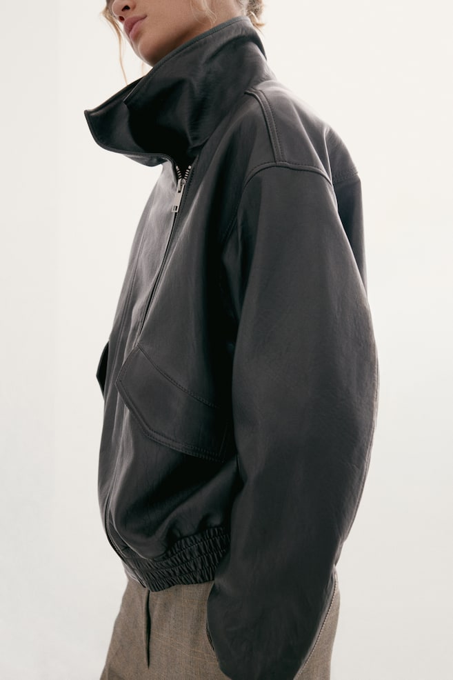 Le mannequin porte une veste en cuir sombre oversize, assortie à un pantalon marron texturé. La veste présente un col cheminée montant, une fermeture zippée asymétrique sur le devant, des épaules tombantes, une poche à rabat à lâavant et un ourlet élastiqué smocké. - 1