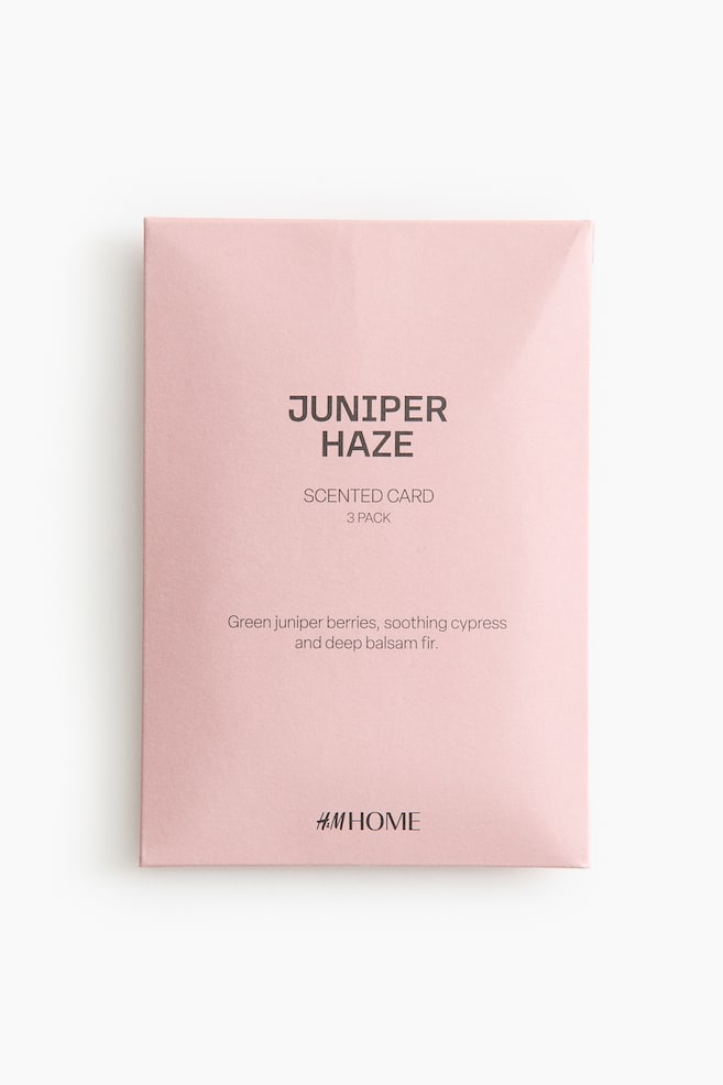 Un paquet rectangulaire rose clair pour "Scented Card 3 Pack" est orienté vers l'avant, affichant en évidence "JUNIPER HAZE" en haut. Il montre également les notes de parfum de baies de genévrier vertes, de cyprès apaisant et de sapin baumier profond, avec le logo H&M HOME apparaissant en bas. - 1