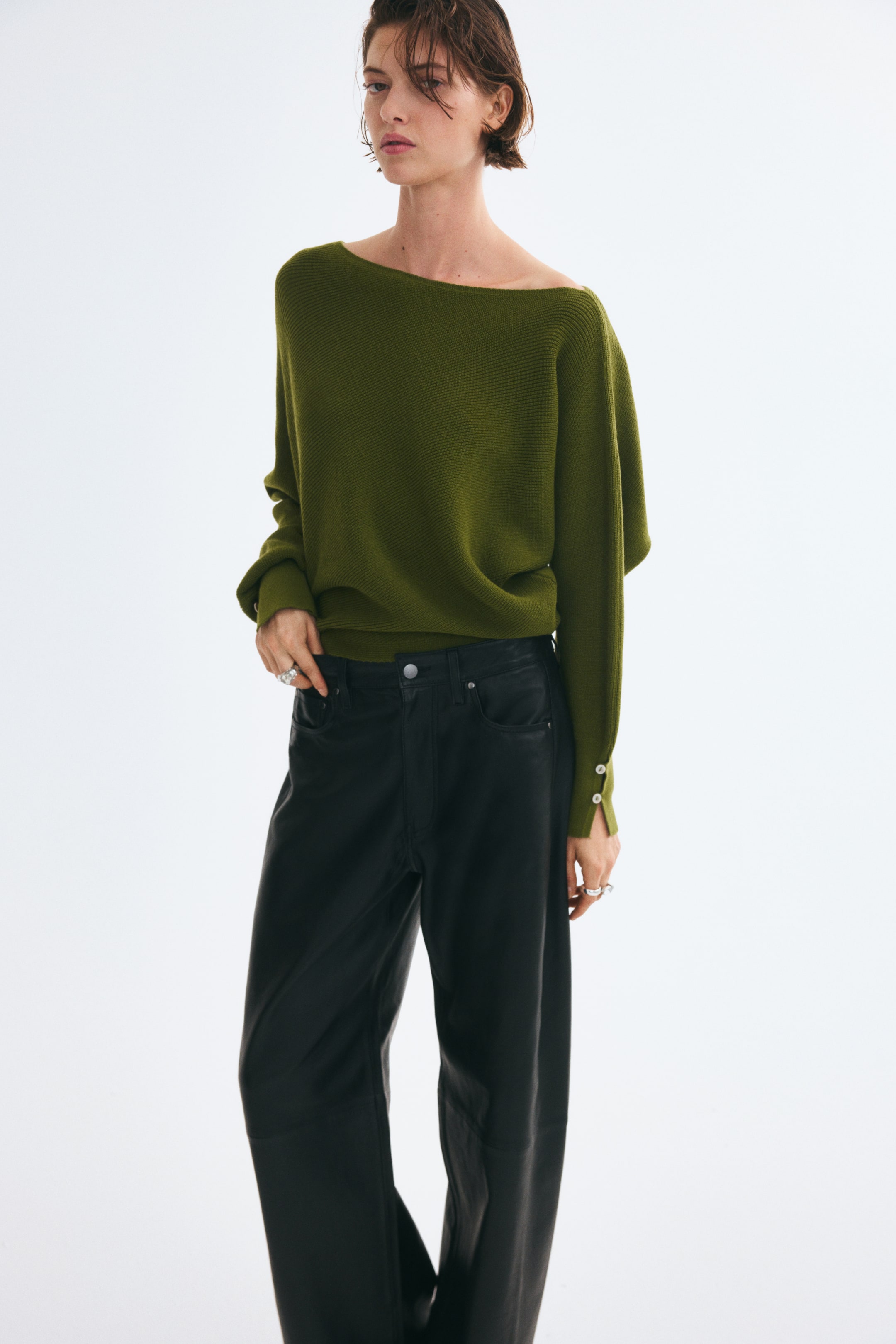 Ver imagen más grande: La modelo lleva un jersey de punto verde oliva con escote asimétrico y mangas largas de murciélago con puños de botón. El jersey de corte holgado se combina con un pantalón negro de pata ancha.