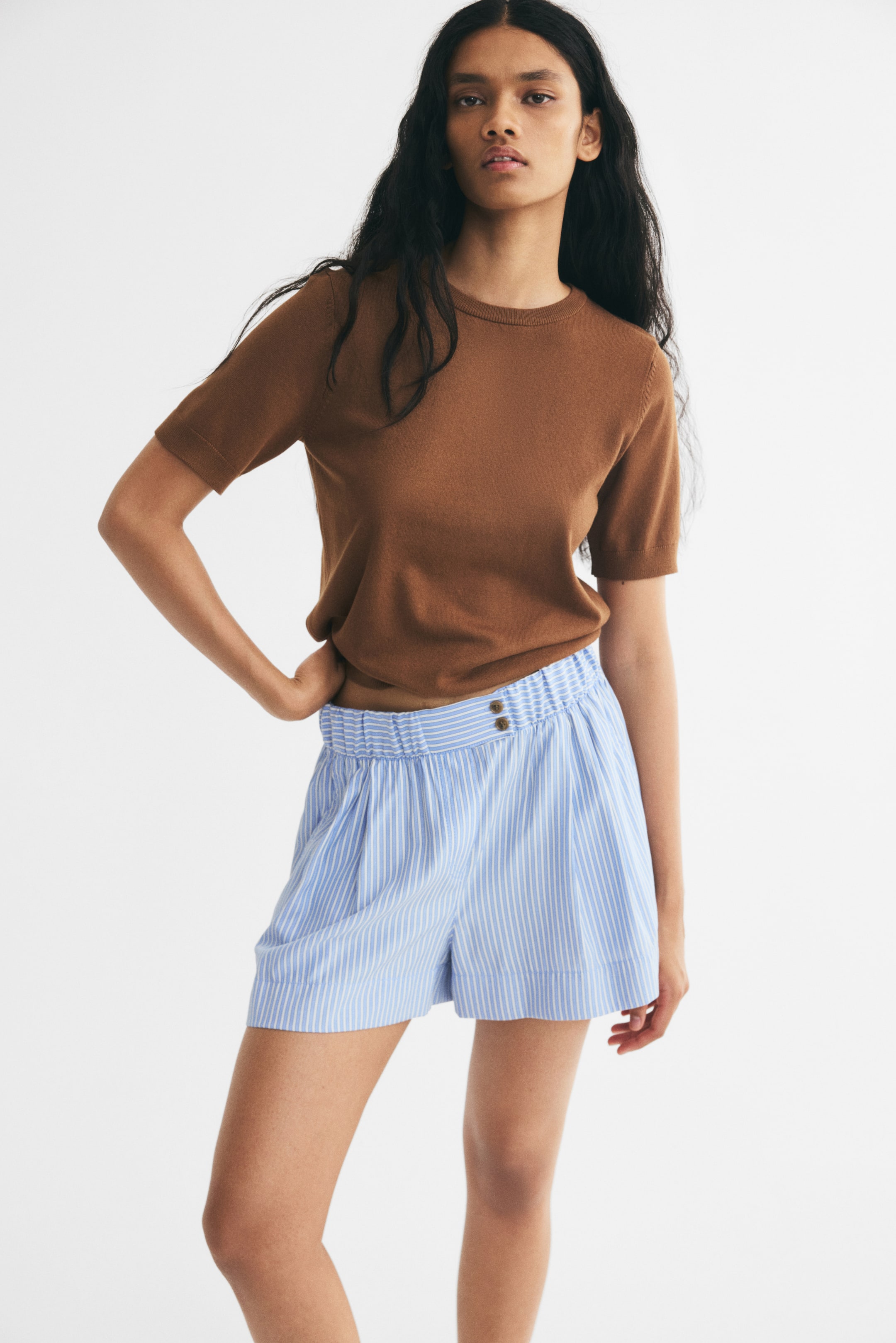 Ampliar la imagen: La modelo lleva una blusa de tejido de manga corta color café, metida dentro de unos shorts a rayas azul claro y blanco, con pretina fruncida y elástica, dos botones visibles y pinzas al frente.