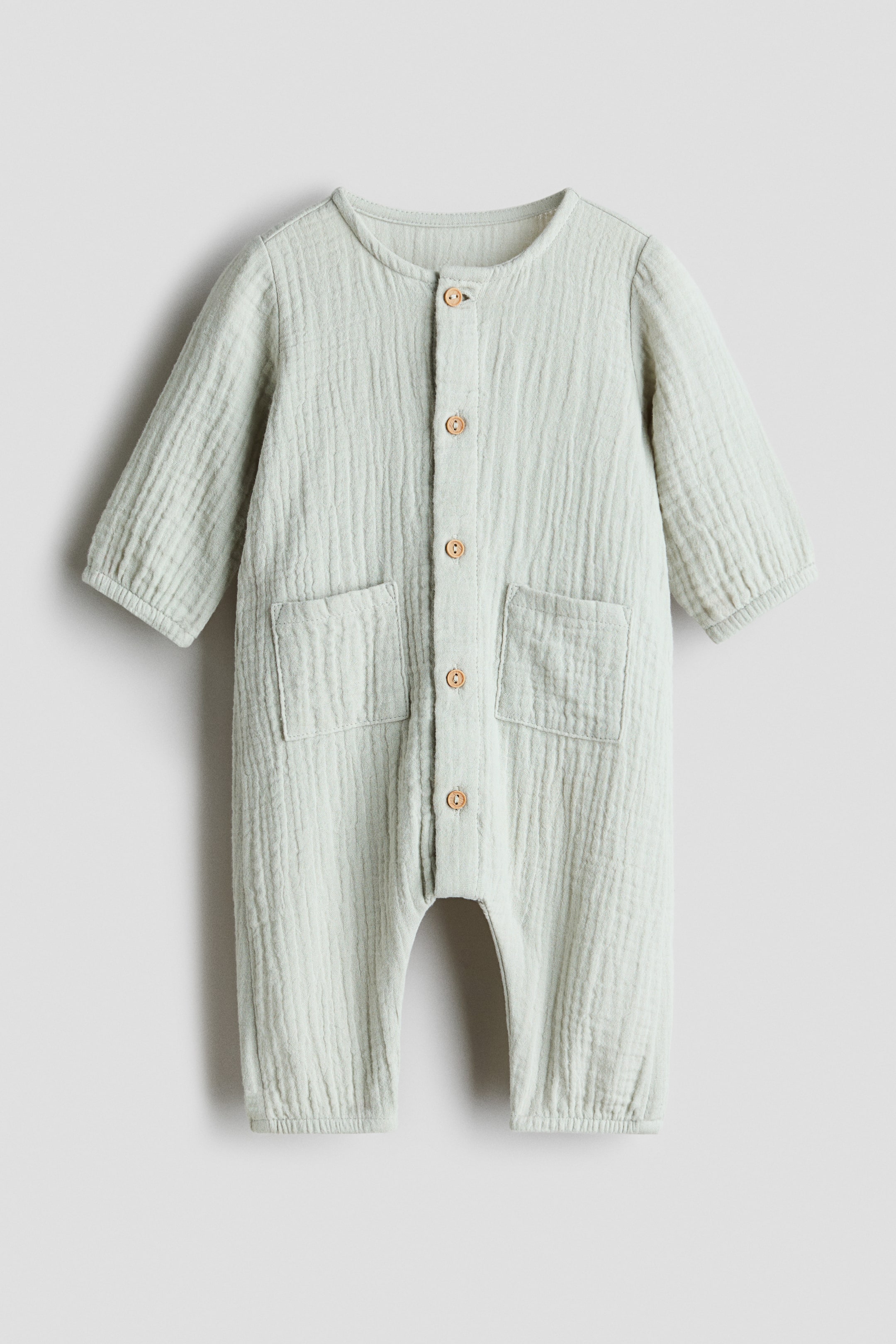 View larger image: A long-sleeved romper suit in light green soft cotton muslin is presenting its FRONT. It is featuring a round neckline, a button-down front with natural-colored buttons, and two patch pockets. Covered elastication is gathering the cuffs and leg hems.