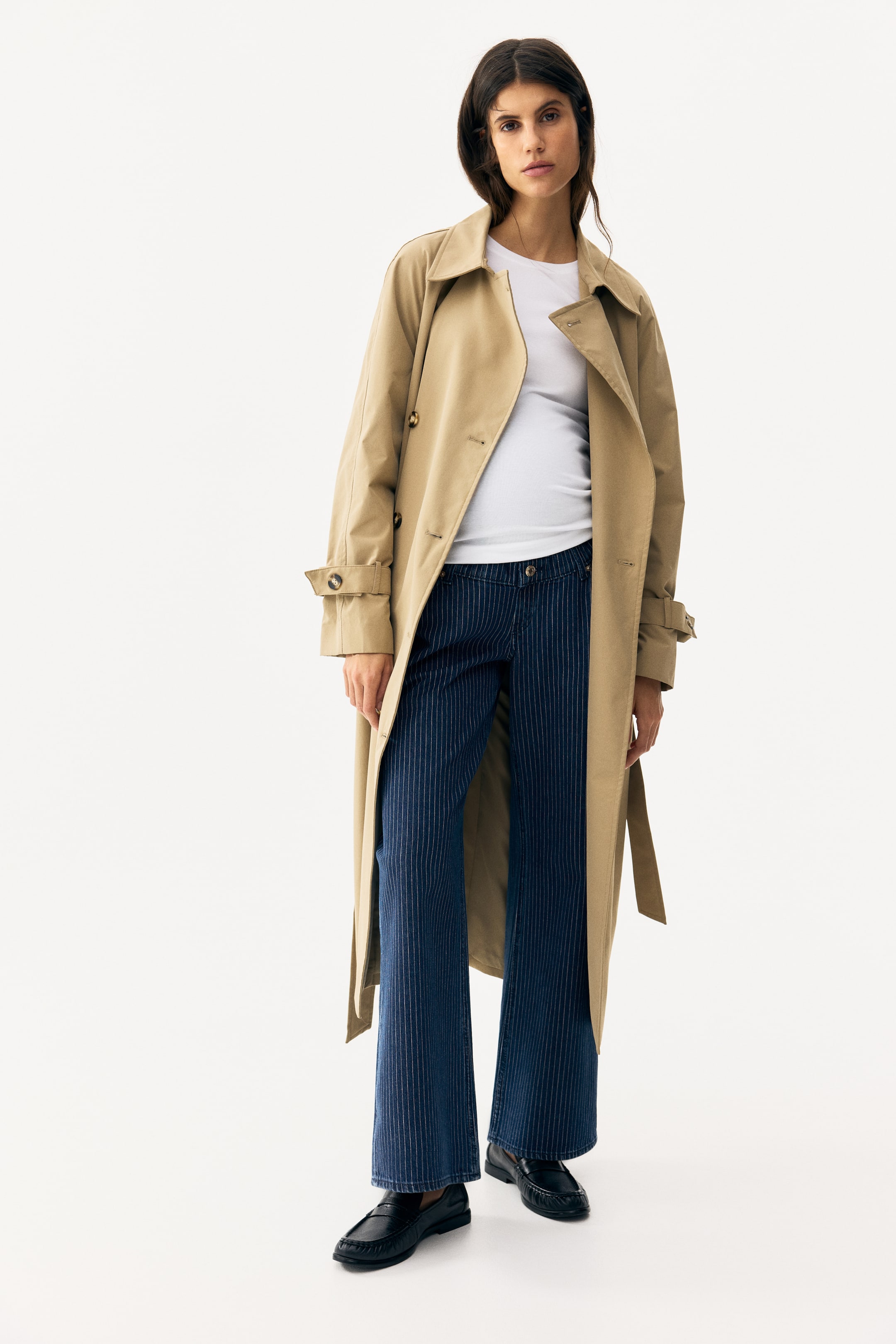 Visualizza immagine più grande: Un trench coat color cammello viene presentato frontalmente, leggermente inclinato verso destra, indossato aperto sopra una maglietta bianca. Presenta un classico colletto a punta, uno stile doppiopetto con bottoni visibili sulla paramontura destra, spalline sulle spalle e cinturini regolabili sui polsini. Una cintura in tessuto coordinata è appesa non allacciata sul lato.