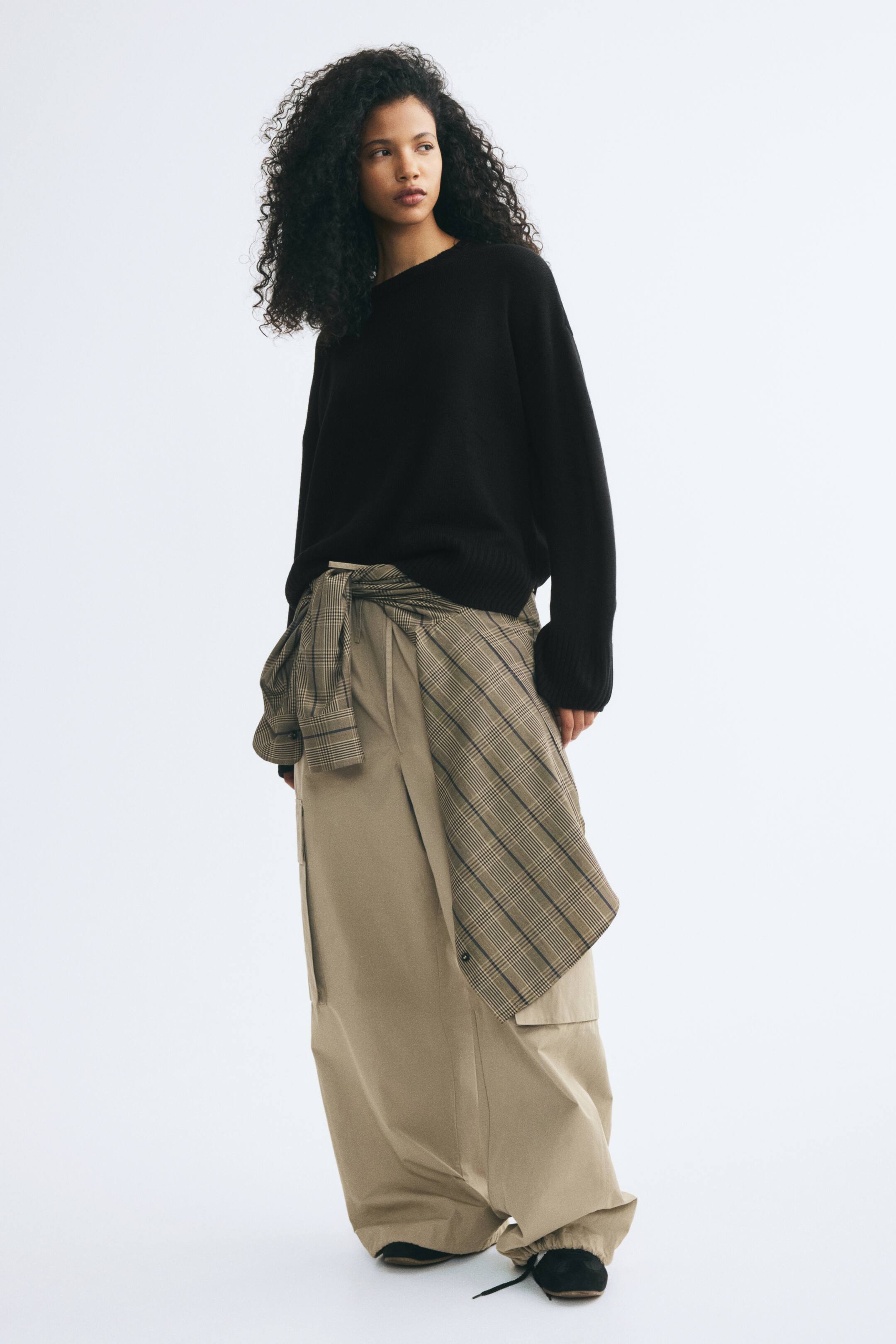 Ver imagen más grande: La modelo lleva un jersey de punto negro de corte relajado, con cuello redondo y hombros caídos. El jersey presenta puños acanalados y se combina con pantalones anchos beige y una camisa de cuadros anudada a la cintura.