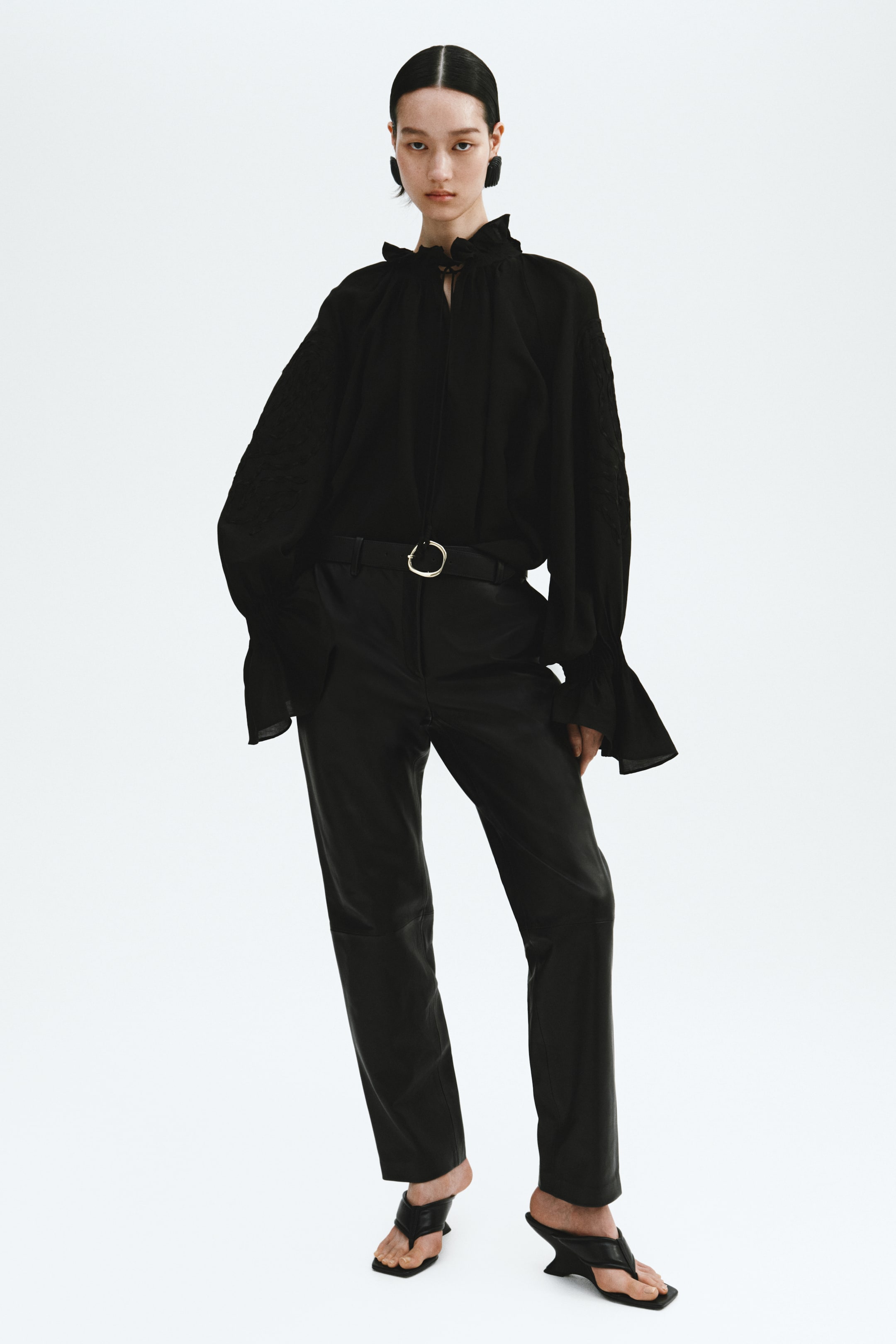 Grotere afbeelding bekijken: Een oversized zwarte blouse in een luchtige weefsel wordt gedragen door het model, ingestopt in een zwarte broek. Het heeft een geplooide opstaande kraag, een V-vormige opening met smalle strikjes, extra lange raglanmouwen met ton-sur-ton geborduurde details en geplooide, subtiel uitlopende manchetten.
