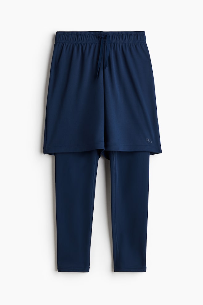 A pair of navy blue sports shorts is being layered over navy blue jersey tights, presented from the front. The shorts are featuring an elasticated, drawstring waist, and a small reflective symbol is appearing on the lower right leg. - 1
