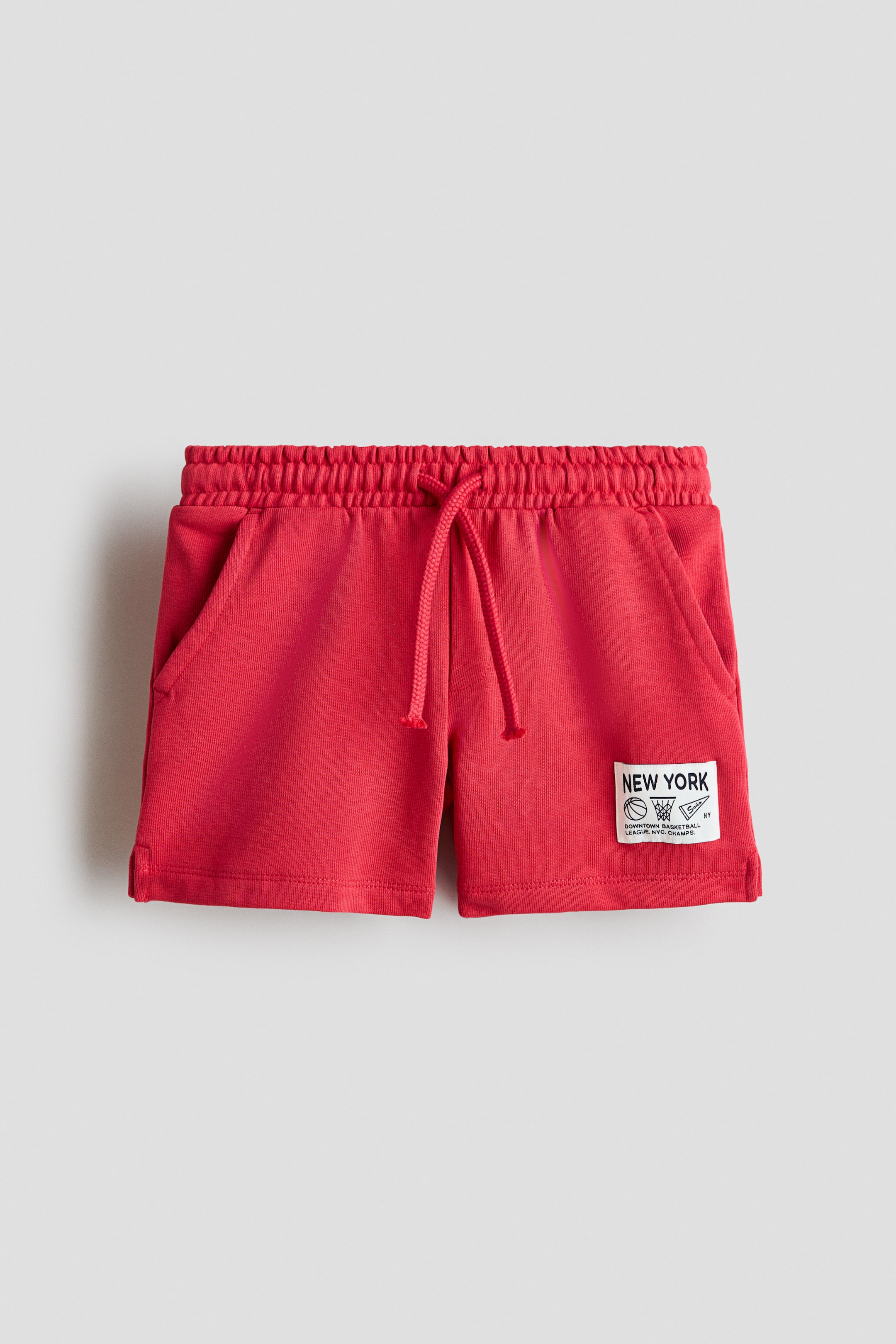 View larger image: Red shorts come with an elasticated waistband featuring a red drawstring tie and two slanted front pockets. A white rectangular patch is stitched onto the lower right leg, showcasing the "NEW YORK" text, basketball-themed icons, and the "DOWNTOWN BASKETBALL LEAGUE N.Y.C. CHAMPS" text.