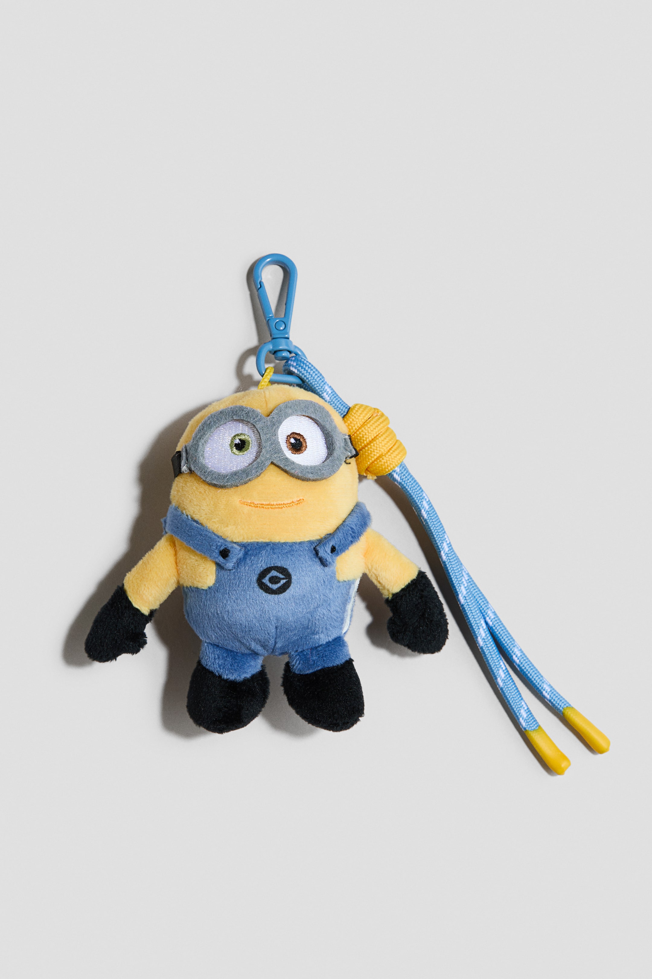 Ver imagen más grande: Un llavero con un personaje de peluche Minion está colgado y mirando hacia adelante, mostrando un cuerpo amarillo, un peto azul, guantes y pies negros, y gafas grises con un ojo verde y otro marrón. Un mosquetón azul conecta el llavero a un cordón trenzado azul y blanco que termina en puntas de plástico amarillo.