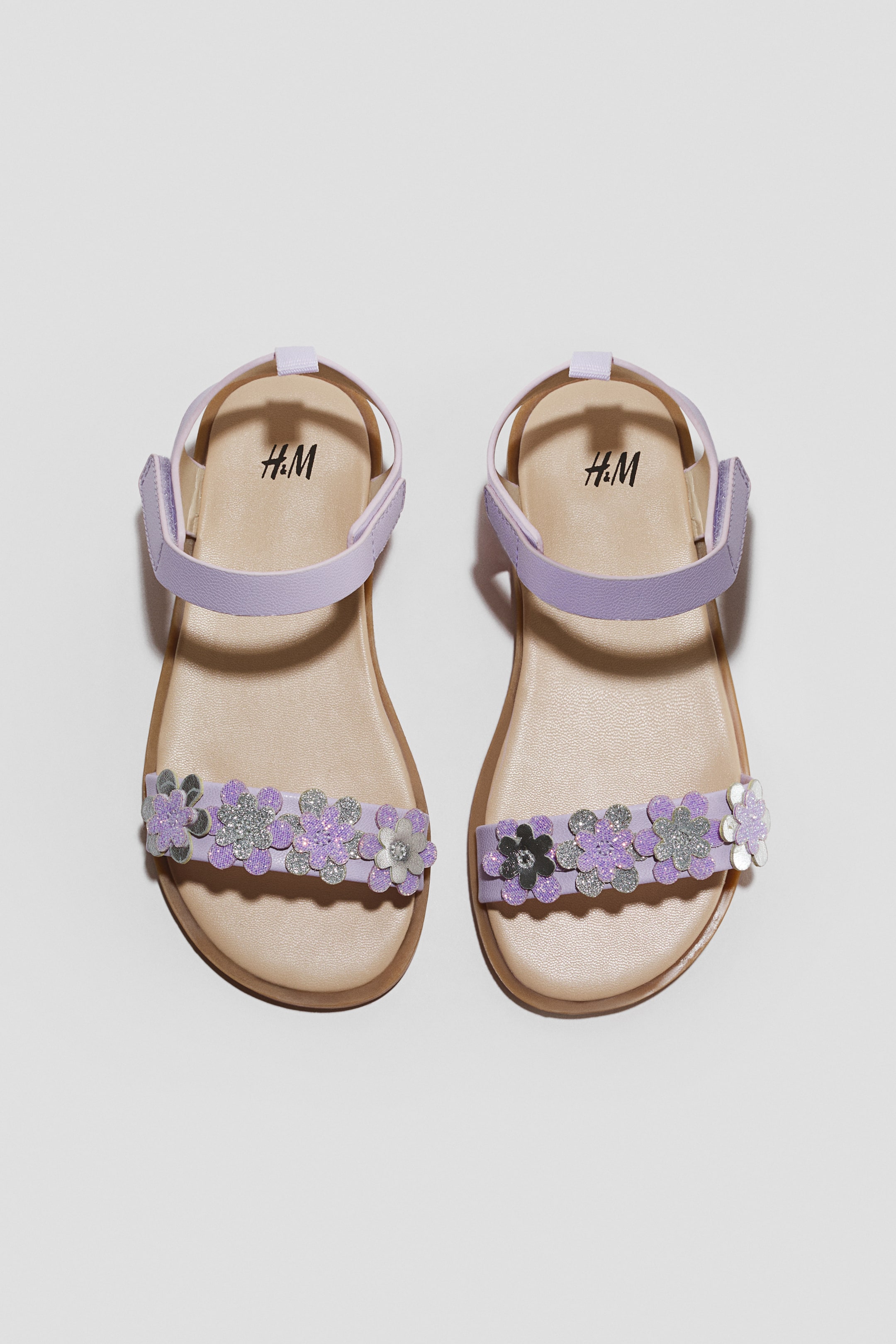 View larger image: These light purple children’s sandals are shown from a top-down view, featuring flat beige footbeds and thin brown soles. The front toe straps are decorated with a row of glittery flower appliqués in shades of purple and silver, while the ankle straps feature adjustable hook-and-loop fasteners.