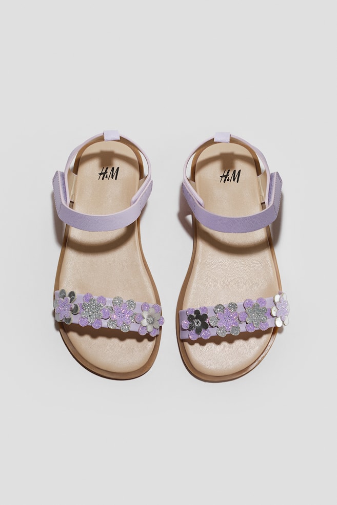 These light purple childrenâs sandals are shown from a top-down view, featuring flat beige footbeds and thin brown soles. The front toe straps are decorated with a row of glittery flower appliquÃ©s in shades of purple and silver, while the ankle straps feature adjustable hook-and-loop fasteners. - 1
