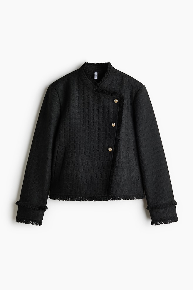 A black textured tweed jacket is facing front, featuring an asymmetrical closure secured by three gold-tone buttons and a standing collar. The jacket is detailed with frayed fringe trim along the collar, front opening, cuffs, and hem, and includes subtle side welt pockets. - 1