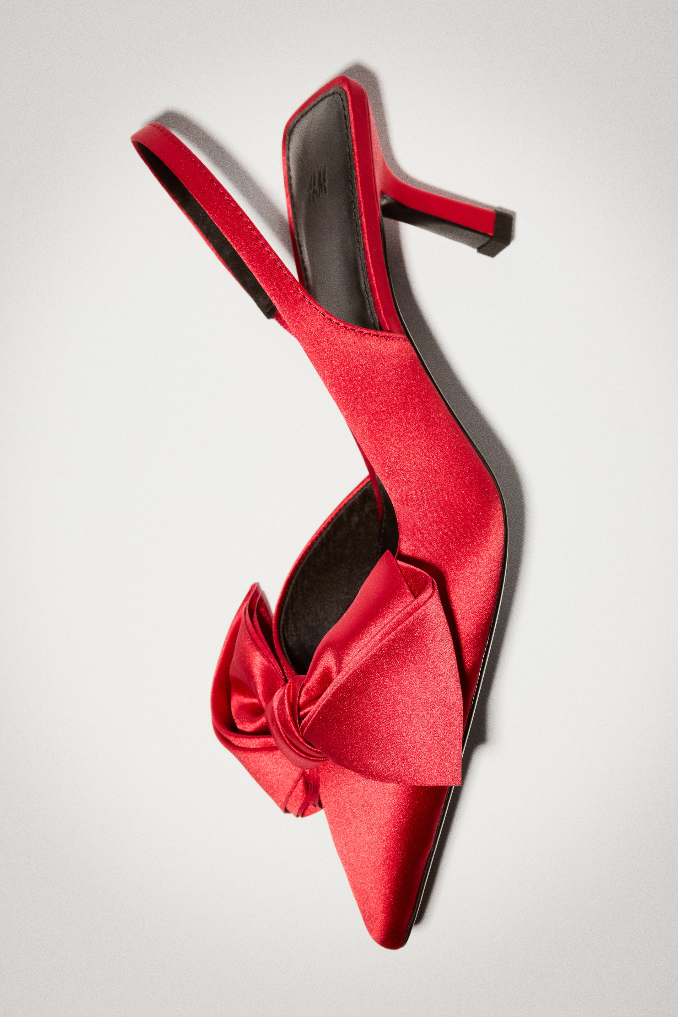 View larger image: A red satin slingback heel showcases a pointed toe with a large decorative bow at the front. The shoe features a matching elasticized slingback strap, a black insole, and a partially covered stiletto heel.
