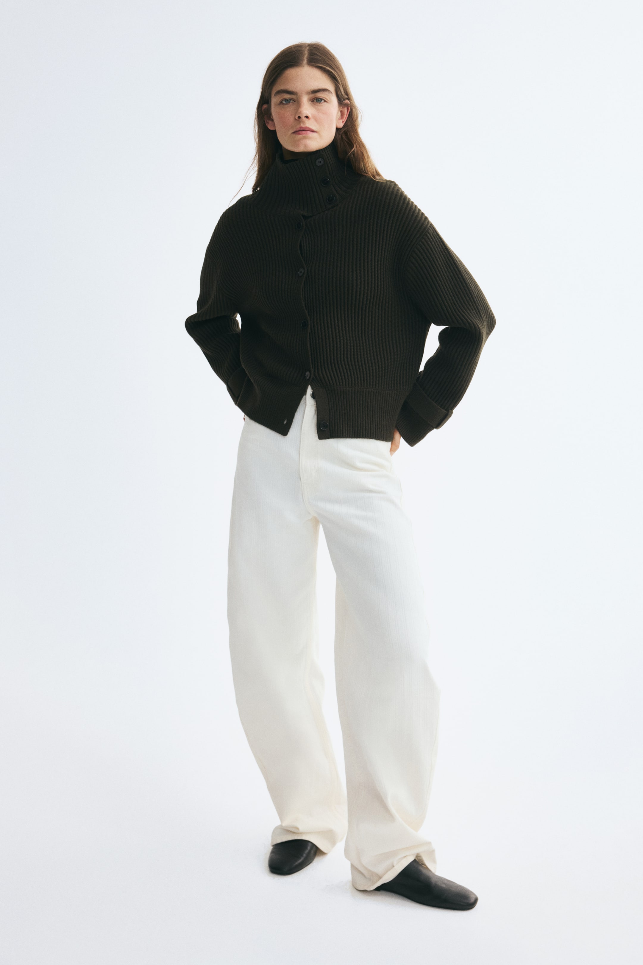 Agrandir l'image: Un cardigan côtelé brun foncé, doté d’un col montant avec patte boutonnée, d’une fermeture boutonnée sur le devant et d’épaules tombantes, est porté par le mannequin. Le cardigan est associé à un pantalon blanc à jambes larges et à des chaussures noires.