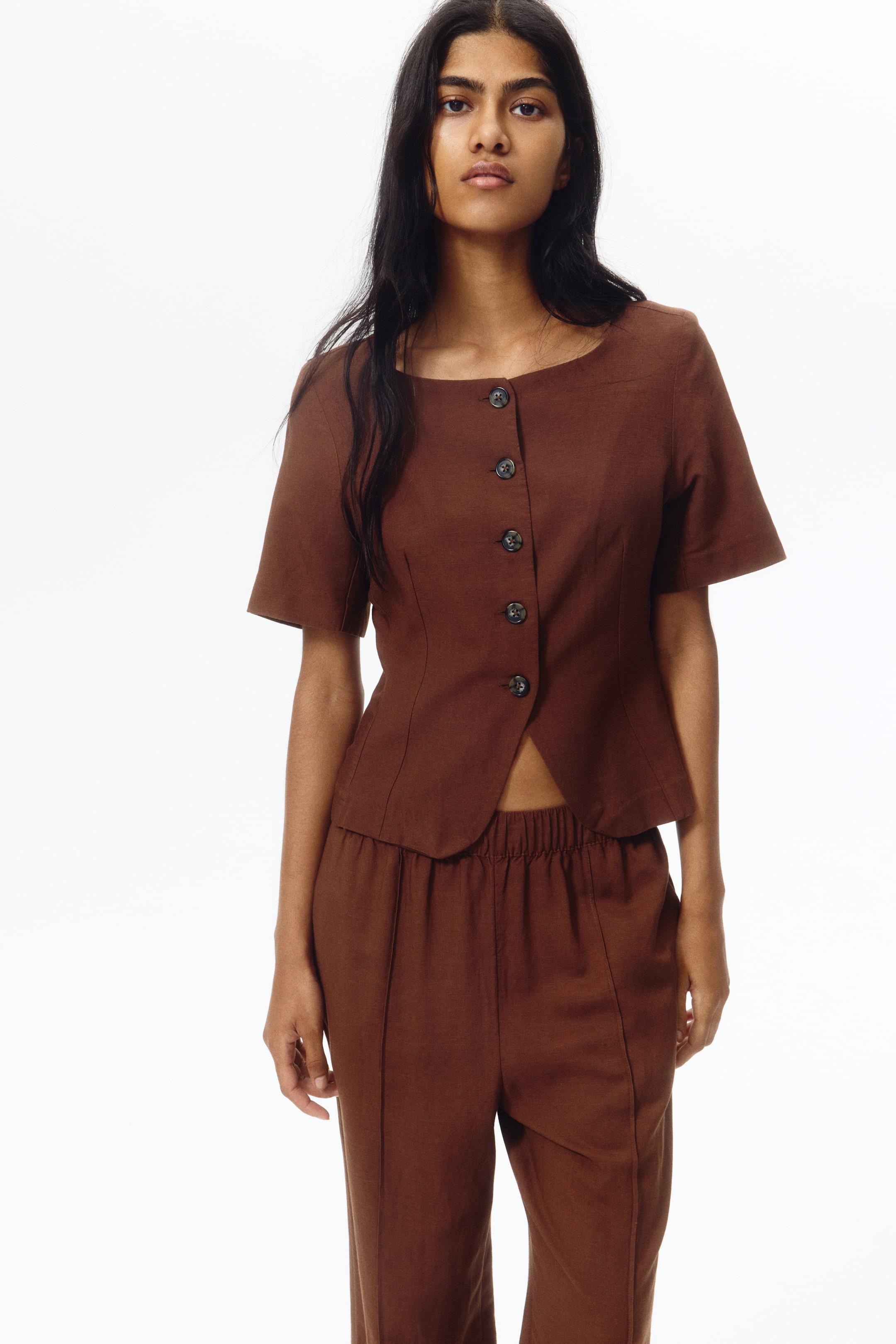 View larger image: Presenting a brown two-piece ensemble, showcasing a short-sleeved top with a broad round neckline, front button fastening, and a gracefully curved cropped hem. The coordinating trousers feature an elasticated waistband and vertical seam accents on the front of each leg.