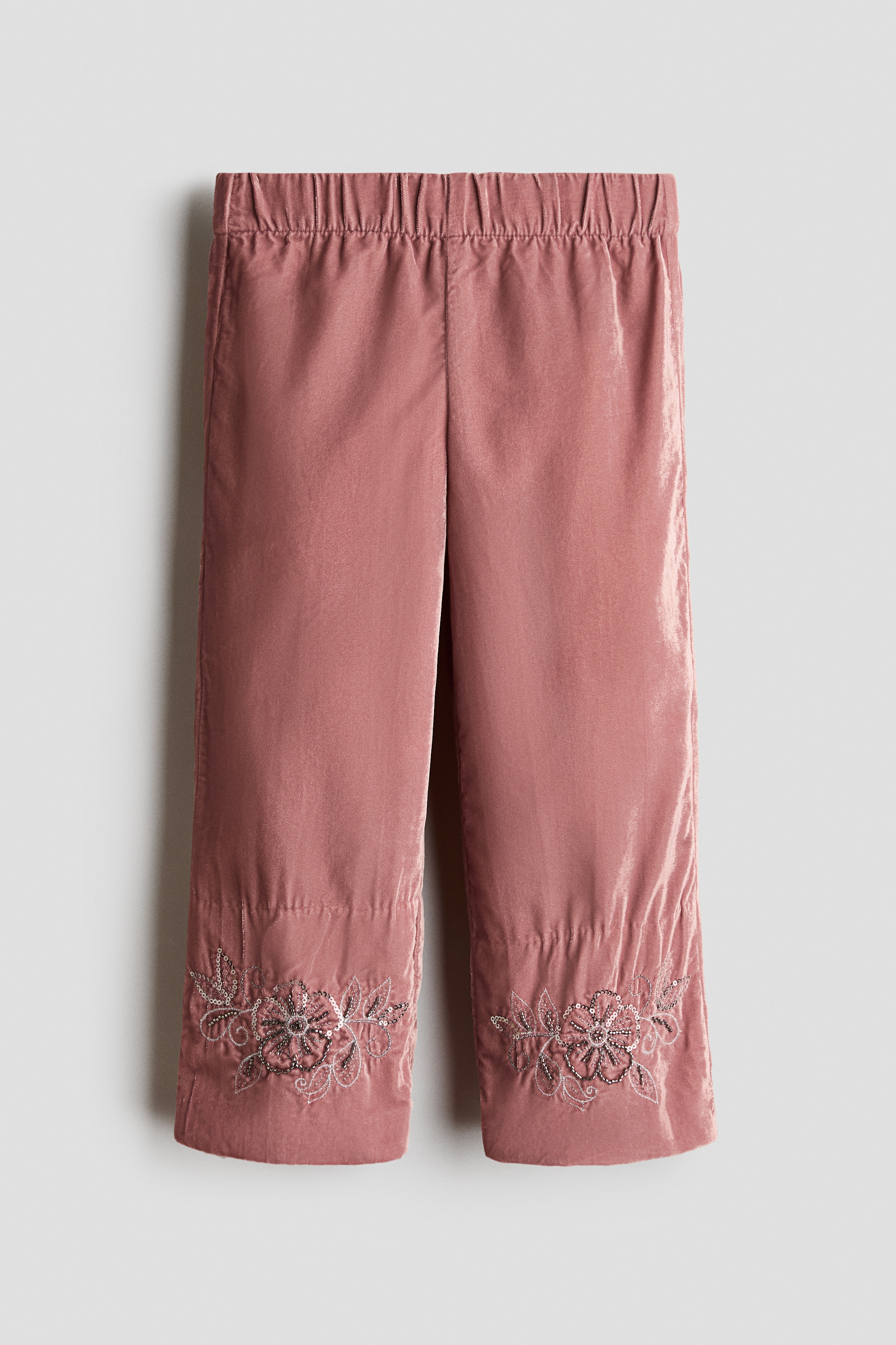 Dusty pink velvet pants are shown from the front, featuring an elasticated waist and straight legs. The pants feature intricate floral embroidery at the hems, detailed with beads and sequins.