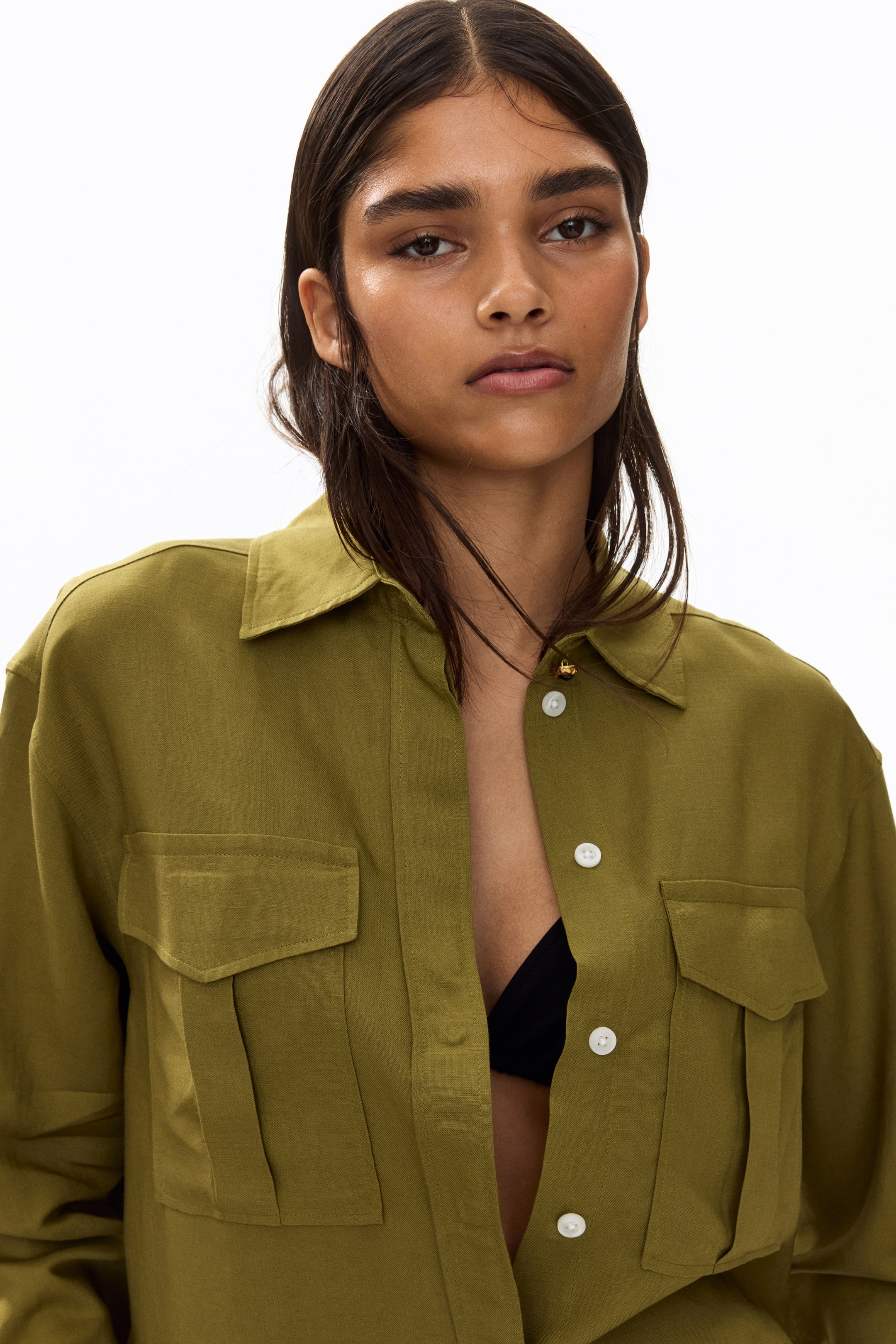 View larger image: An olive green button-down shirt is showcased from the front, highlighting a pointed collar, white buttons, and two flap pockets with pleats on the chest. The front is partially unbuttoned, revealing a black bra underneath.