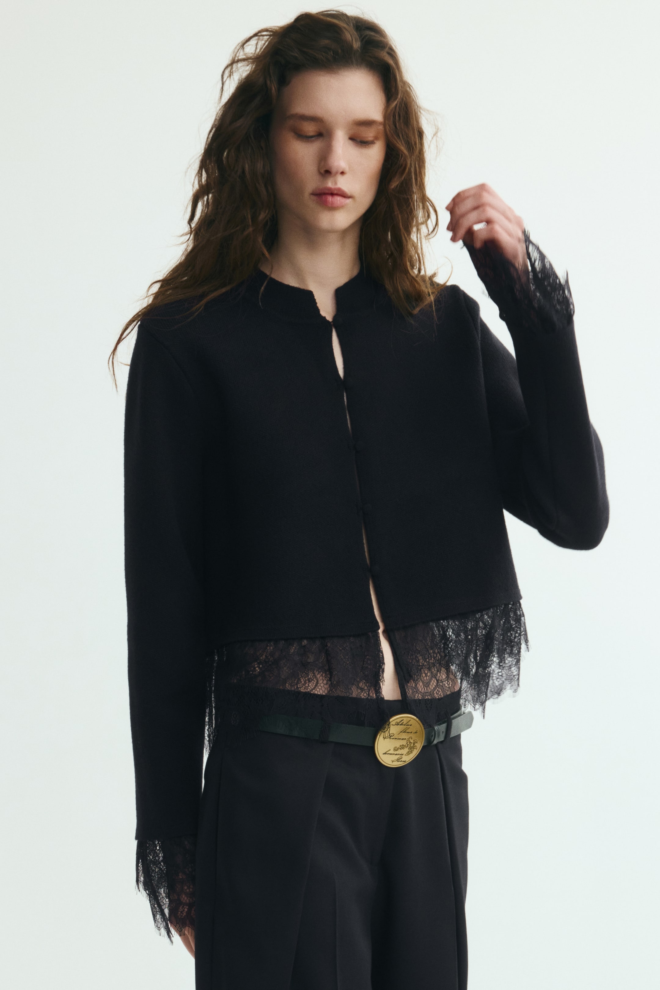 View larger image: The model is wearing a black, cropped, fitted cardigan with a band collar, concealed front buttons, and long sleeves. The cardigan features black eyelash lace trim at the hem and cuffs and is paired with matching black trousers and a belt with a large gold buckle.
