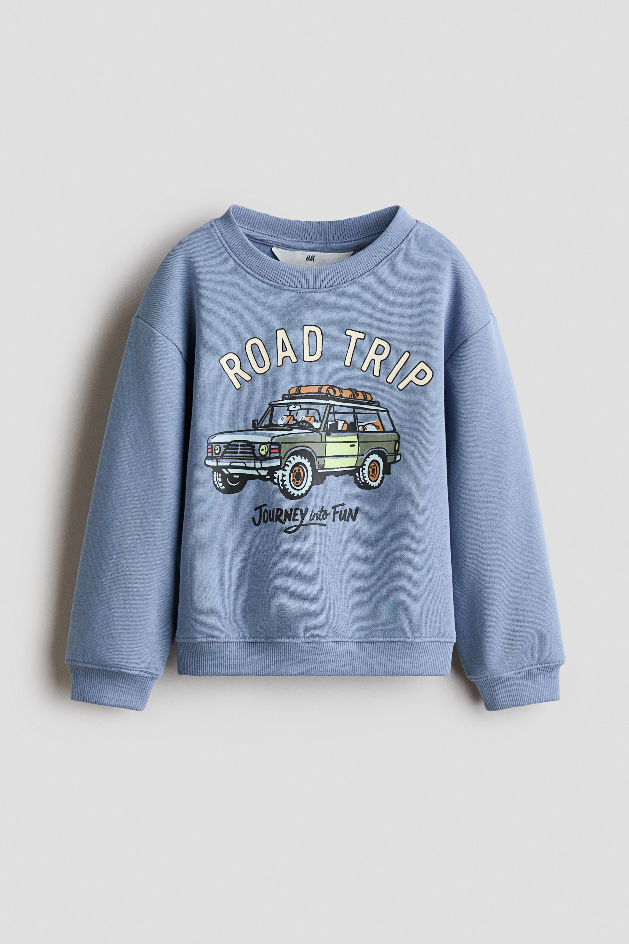Visa större bild: En blå sweatshirt visas framifrån och framhäver en rund halsringning med ribbstickad kant, nedsänkta axlar samt ribbstickade muddar och nederkant. Framsidan visar ett centralt grafiskt tryck av ett grönt terrängfordon med orden "ROAD TRIP" i en båge ovanför och "JOURNEY into FUN" skrivet nedanför.