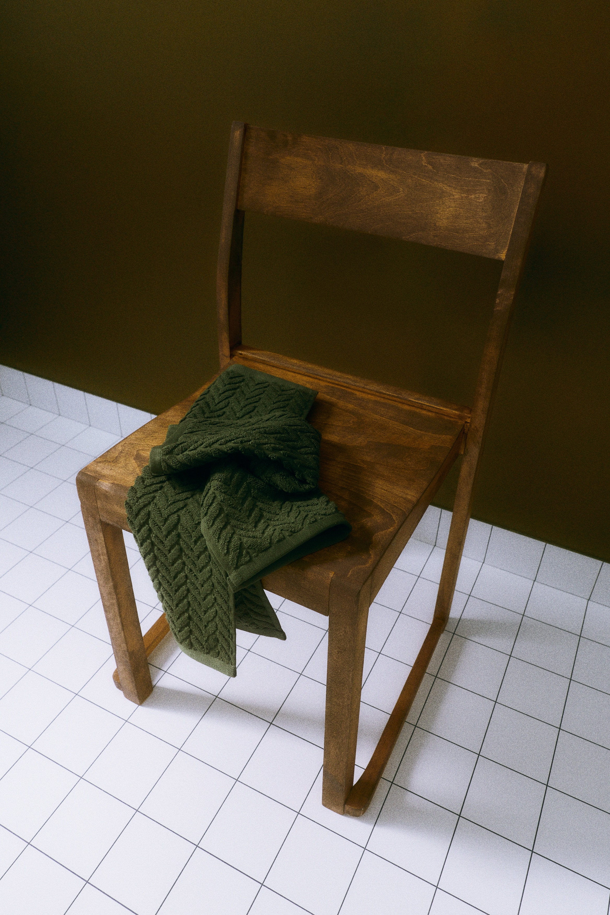 Una coppia di asciugamani da mano in spugna di cotone verde scuro Ã¨ appesa fianco a fianco a un portaoggetti in legno con ganci, mettendo in risalto un motivo chevron testurizzato ispirato alla lavorazione a maglia a trecce. Ogni asciugamano Ã¨ sospeso tramite un passante sul lato corto, evidenziando il tessuto morbido e cadente.