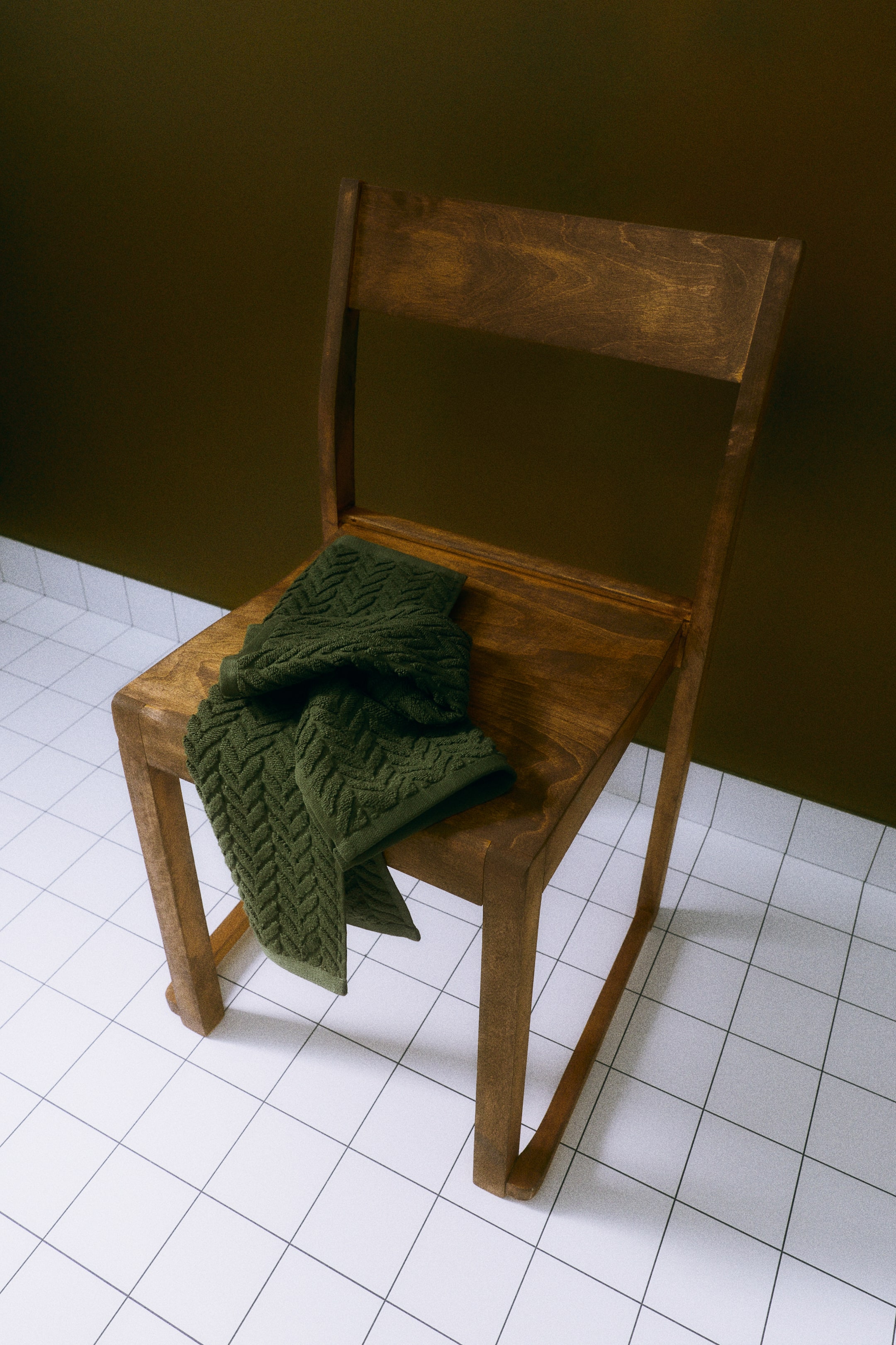 Visa större bild: En mörkgrön handduk i bomullsfrotté är draperad över sitsen på en trästol och framhäver sitt texturerade, kabelstickningsinspirerade chevronmönster.
