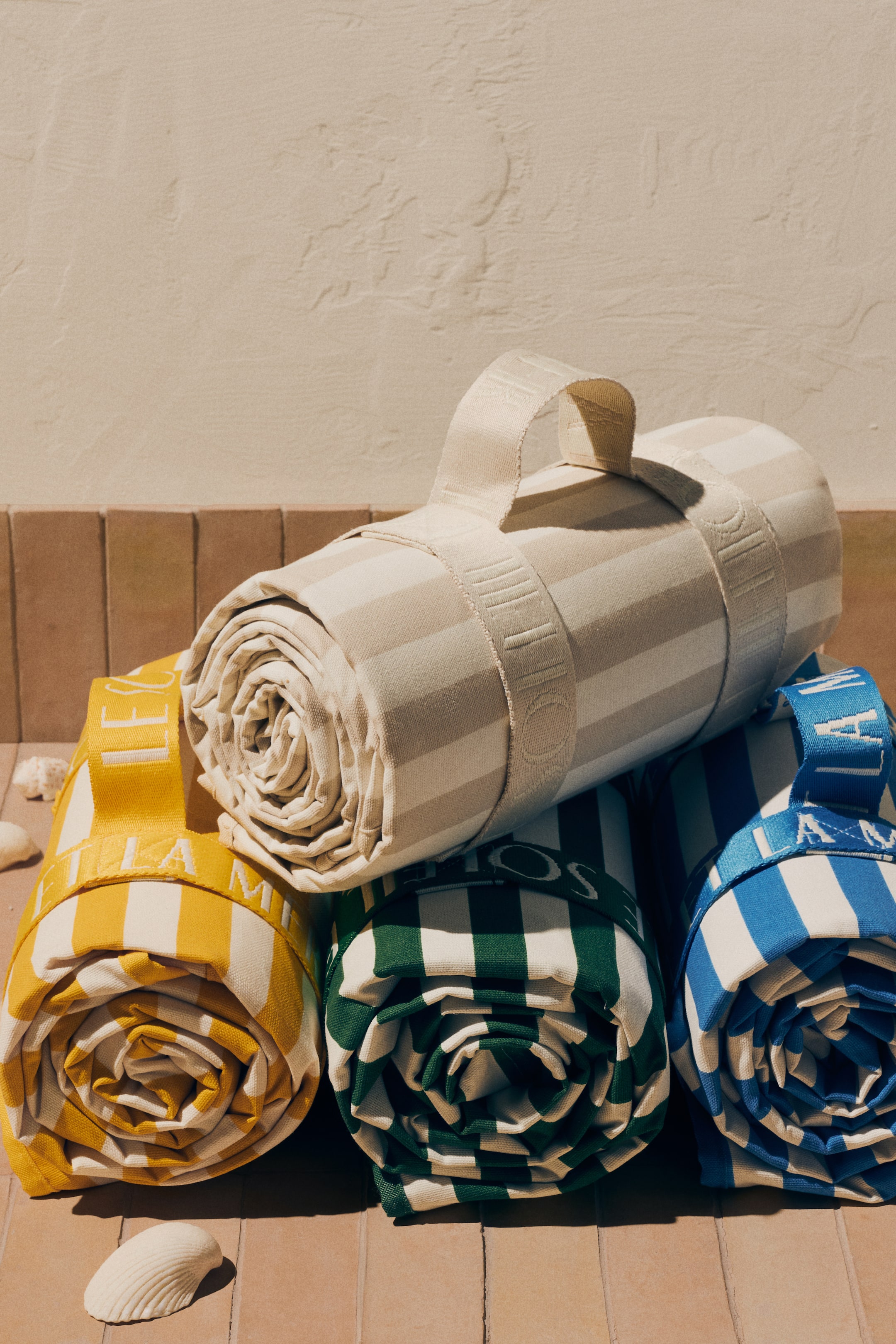 Ampliar la imagen: Cuatro rollos de tela a rayas están apilados, con un rollo beige y blanco colocado horizontalmente sobre tres rollos en amarillo y blanco, verde y blanco, y azul y blanco. Cada rollo está asegurado con una correa de transporte con letras en relieve, y sus extremos enrollados firmemente muestran de manera destacada sus patrones en espiral.