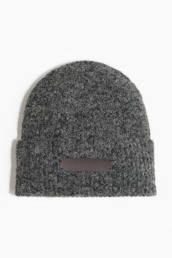 A dark gray beanie is appearing from the front, revealing a textured, heathered knit, with a rectangular dark brown patch being affixed to the front of its folded brim by visible stitching. - 1