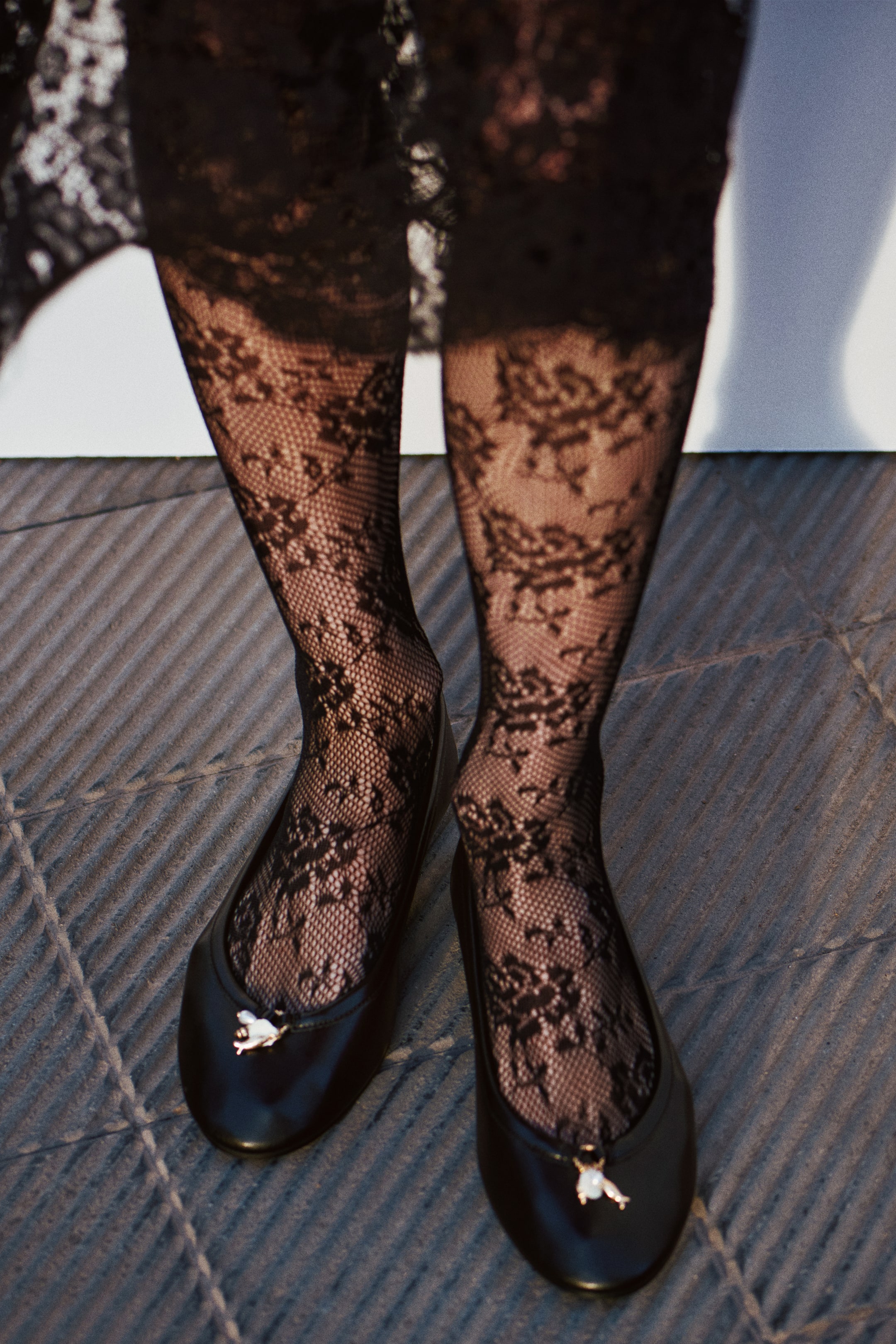 View larger image: Black lace tights, showcasing a detailed floral design, fit snugly on the legs, which are also wearing black ballet flats. Each flat is embellished with a small, light-colored charm on the toe, all captured from a slightly elevated, front-facing angle.