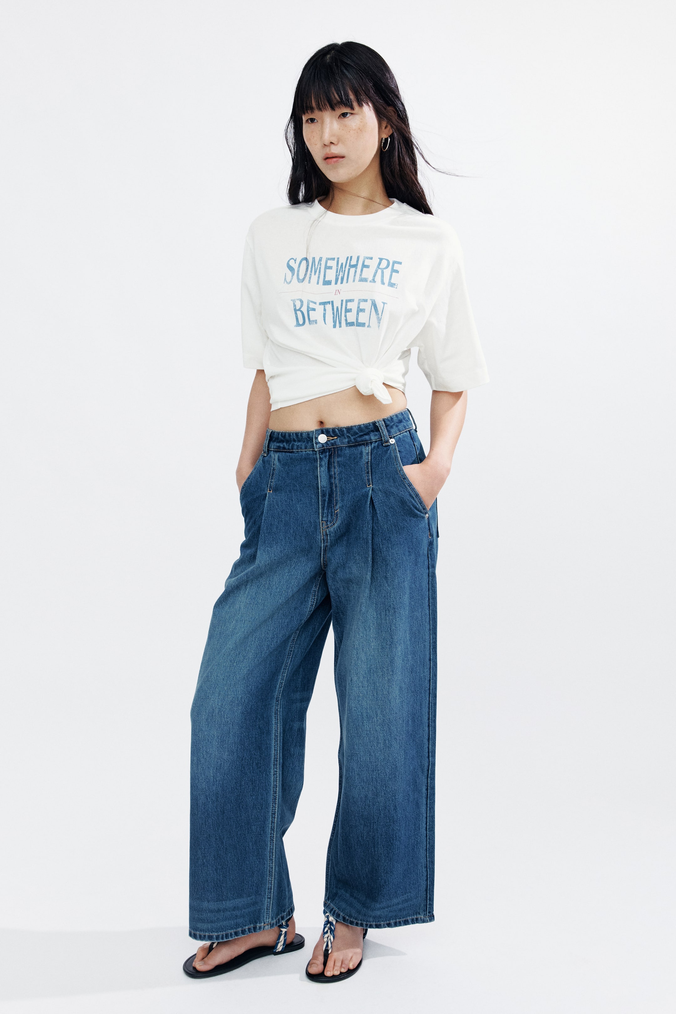 拡大画像を表示: モデルが着用しているのは、ソフトなコットンジャージー素材のボクシースタイルのTシャツで、ブルーの「SOMEWHERE IN BETWEEN」プリントが施されています。リブ付きのネックラインとドロップショルダーが特徴のルーズフィットTシャツは、フロントで結び目を作り、モデルのミッドリフを露出させ、ワイドレッグジーンズと合わせてスタイリングされています。