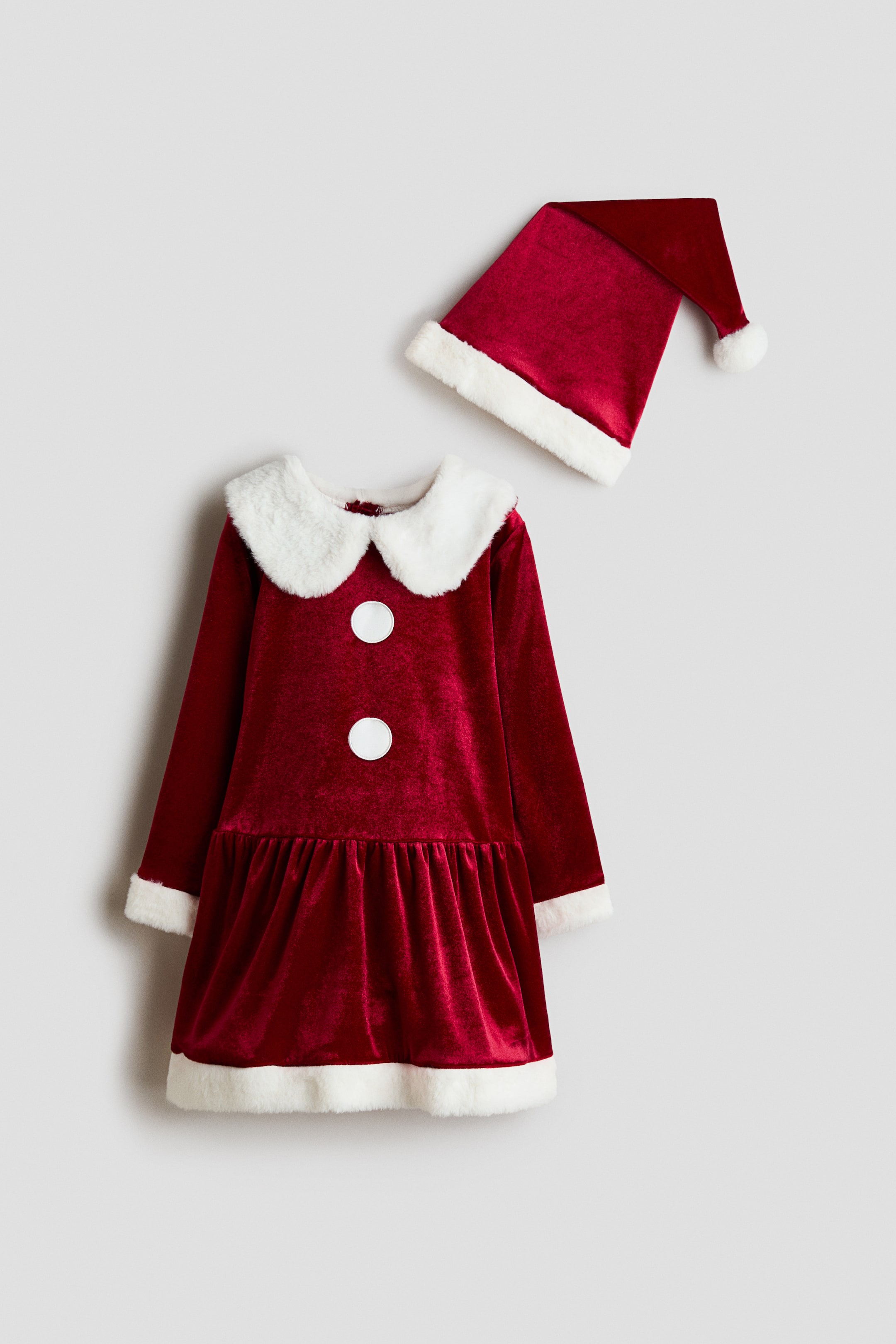 Größeres Bild ansehen: Ein festliches Kinderoutfit besteht aus einem roten langärmeligen Samtkleid und einem passenden spitzen Hut, beide von vorne gezeigt. Das Kleid verfügt über einen weißen Peter-Pan-Kragen aus Kunstfell, eine weiße Kunstfellverzierung an den Ärmelbündchen und am Saum sowie zwei große weiße, runde Details am Oberteil, während der passende Hut mit weißem Kunstfellbesatz und einer Bommel versehen ist.