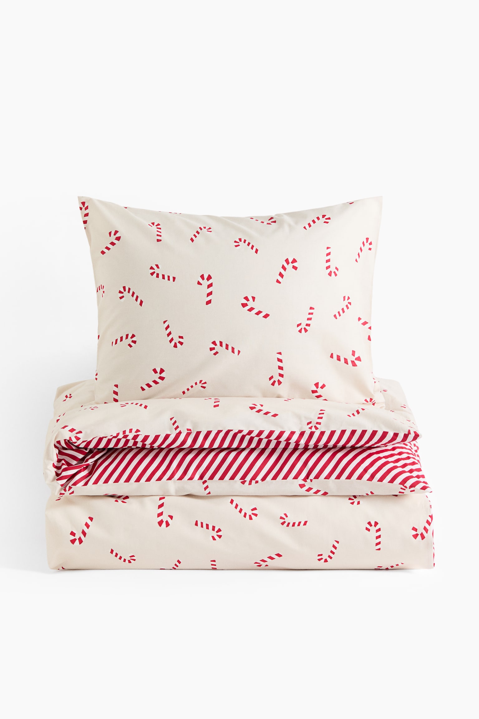 Dieses BettwÃ¤sche-Set, von vorne gezeigt, besteht aus einem cremefarbenen Kissenbezug und einem passenden gefalteten Bettbezug, beide mit einem verstreuten Muster aus kleinen rot-weiÃen Zuckerstangen verziert. Am gefalteten Rand des Bettbezugs ist ein kontrastierendes Futter mit diagonalen roten und weiÃen Streifen sichtbar.