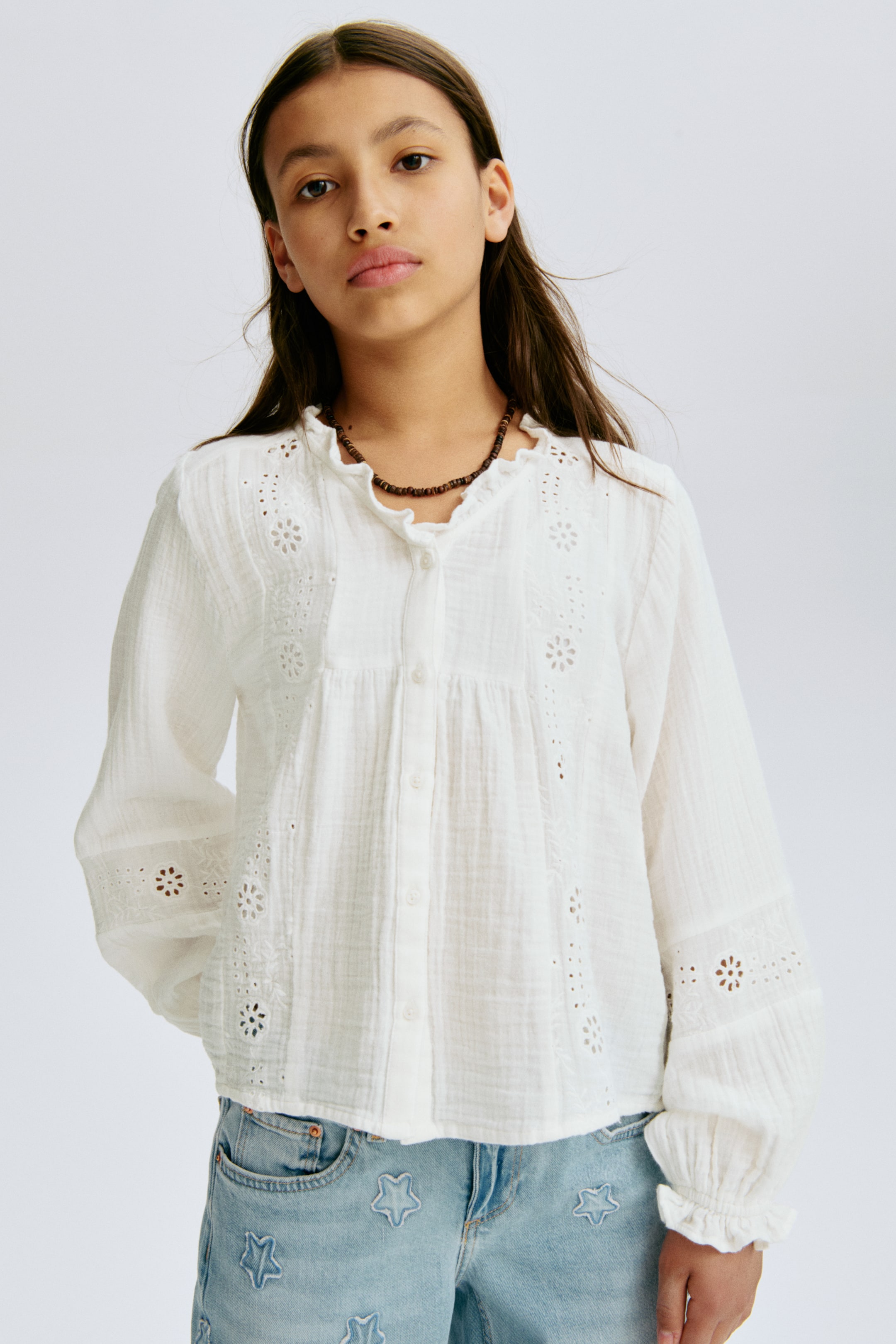 View larger image: The white crinkled cotton blouse features a ruffled stand-up collar and a full button-front placket. It showcases eyelet embroidery details on the gathered front bodice and long sleeves, which end with elasticated ruffled cuffs.