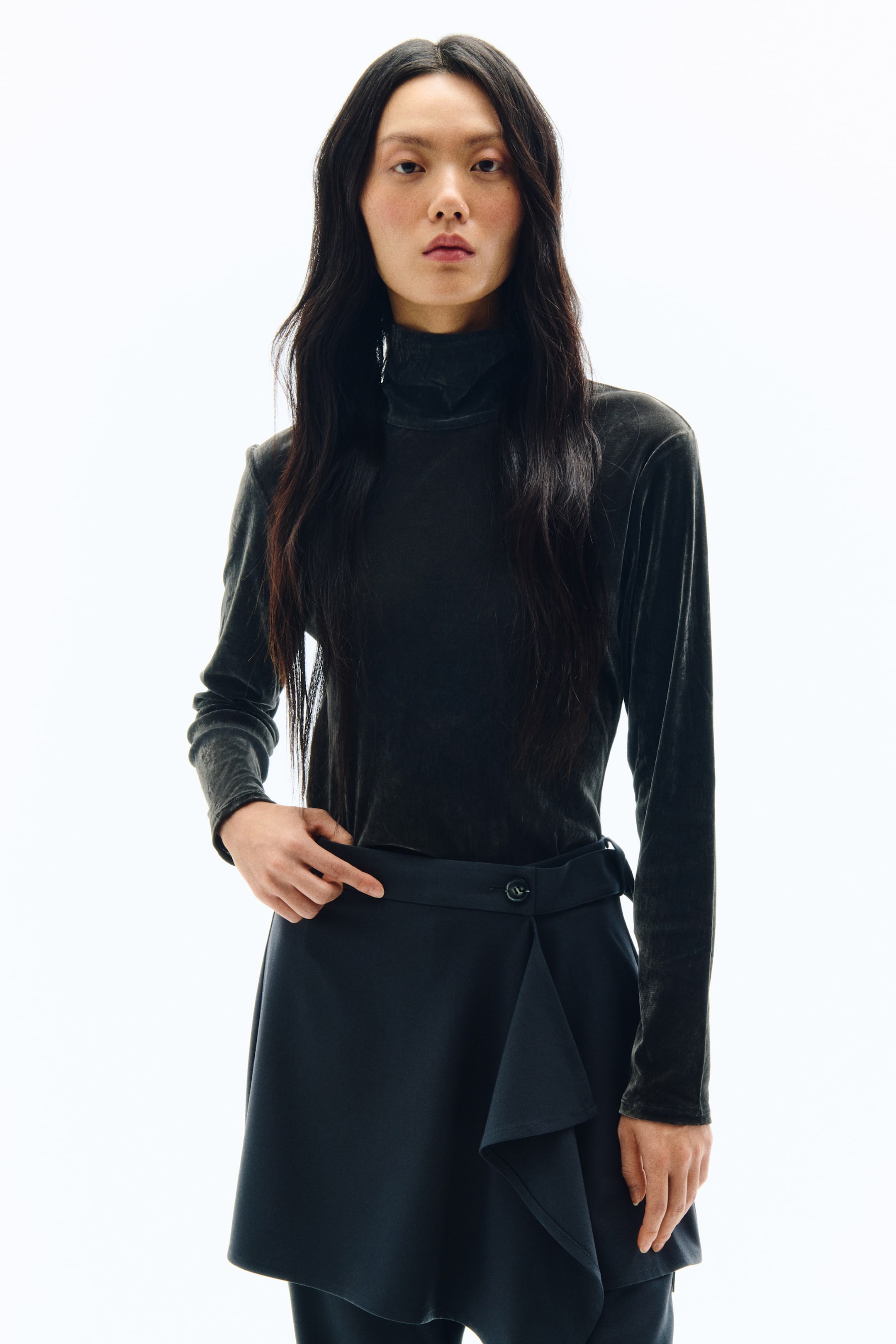 拡大画像を表示: ダークカラーのベルベット調タートルネックトップスを着用しています。ロングスリーブと高めのロールネックが特徴です。合わせているのは、ダークネイビーのラップスタイルスカートで、フロントにアシンメトリーなレイヤードデザインのパネルと、ウエスト部分のボタン留めディテールがあしらわれています。