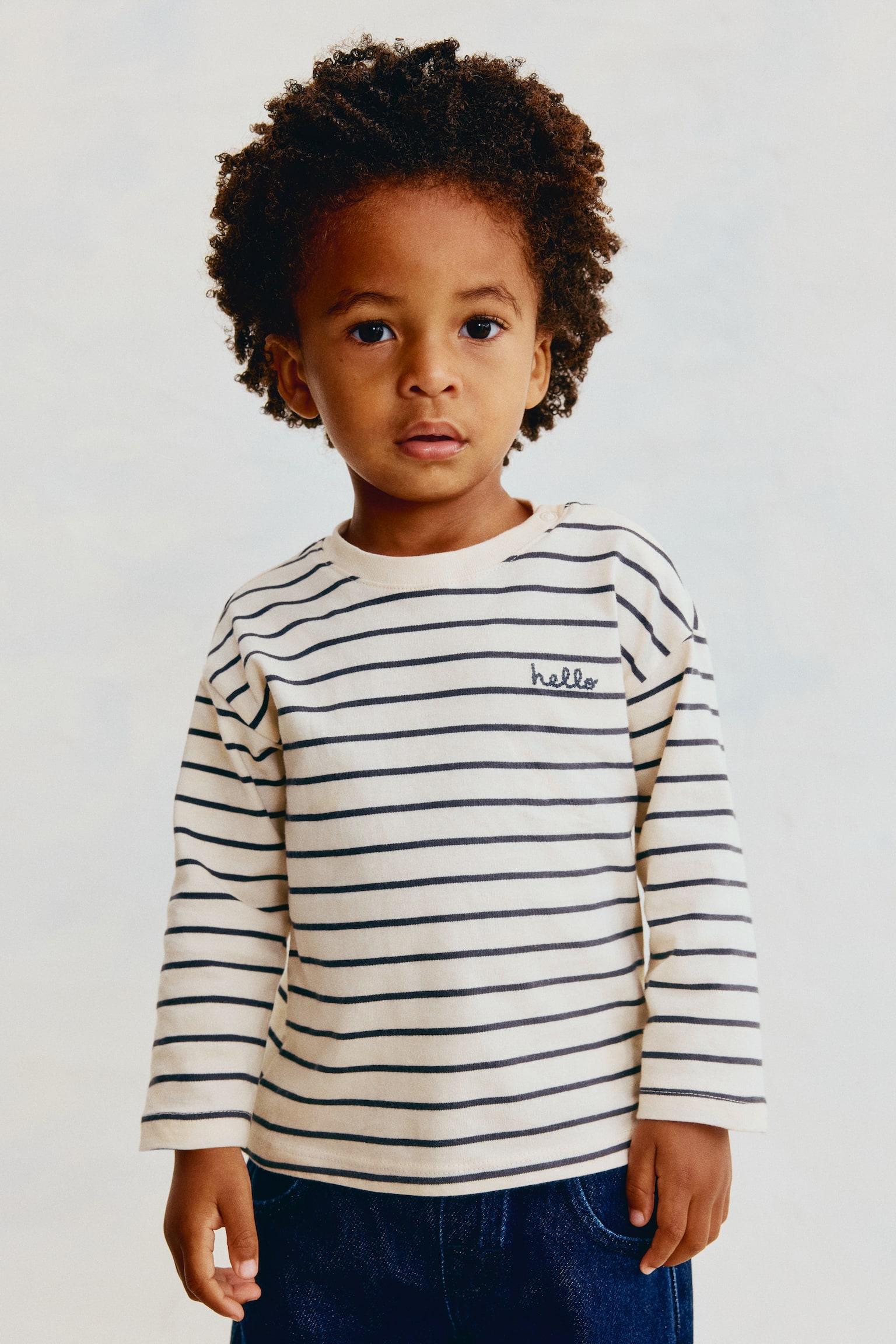 Een kind draagt het H&M Adorables T-shirt met losse pasvorm en lange mouwen, recht van voren afgebeeld om het crÃ¨mekleurige en donker gestreepte patroon te tonen. Het T-shirt heeft verlaagde schouders, een geribde ronde hals met drukknoopsluiting op de schouder en een klein cursief geborduurd "hello" op de linkerborst. - 1