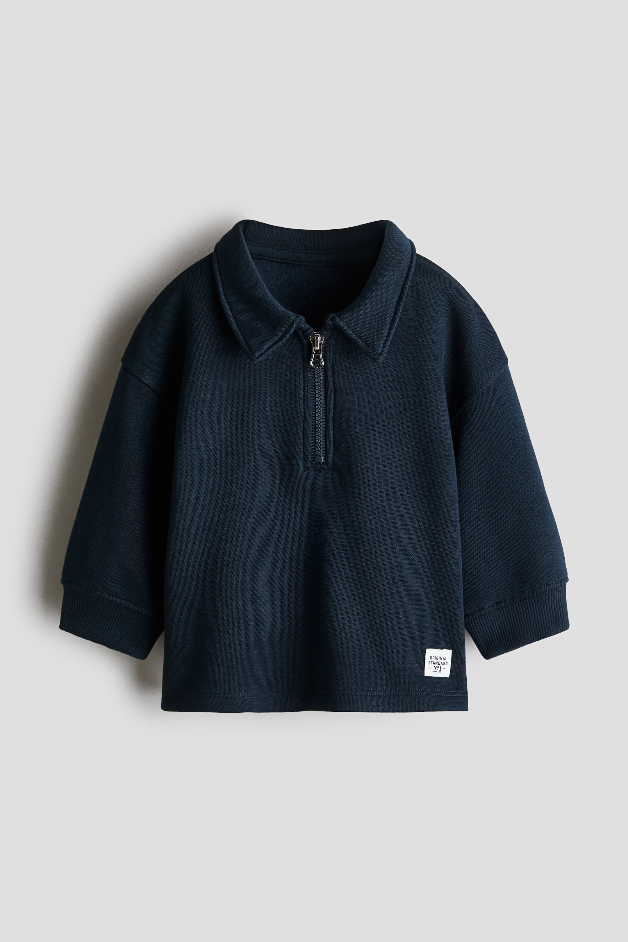 View larger image: A navy blue long-sleeve sweatshirt is facing front, featuring a polo-style collar with a metallic half-zip closure, dropped shoulders, and ribbed cuffs. A small white rectangular brand patch is positioned near the bottom left hem.