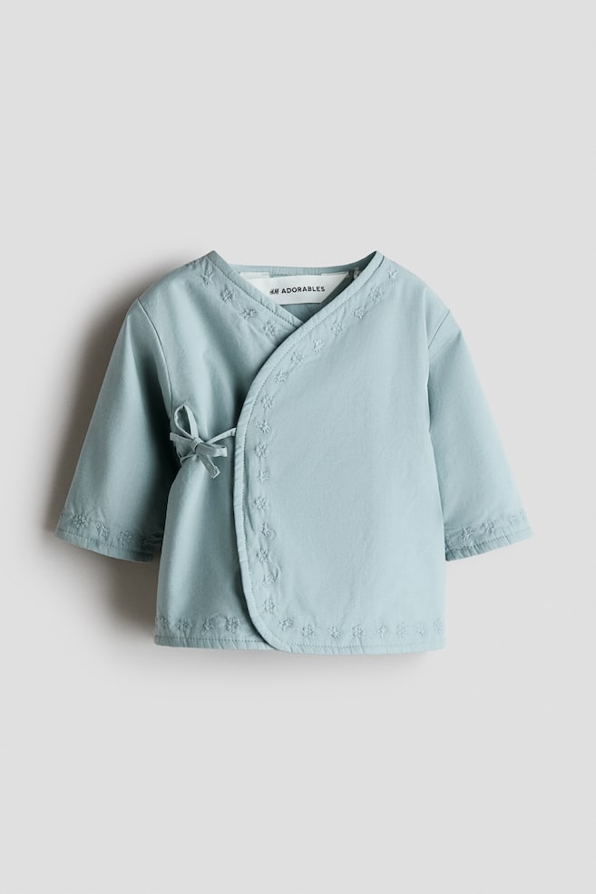 Ein hellblaues Baby-Kimono-Oberteil präsentiert seine Vorderseite mit einem Wickeldesign und einem seitlichen Bindeband, das eine Schleife bildet. Bestickte Motive zieren den V-Ausschnitt, die überlappenden Vorderkanten, die Ärmelbündchen und den unteren Saum. - 1