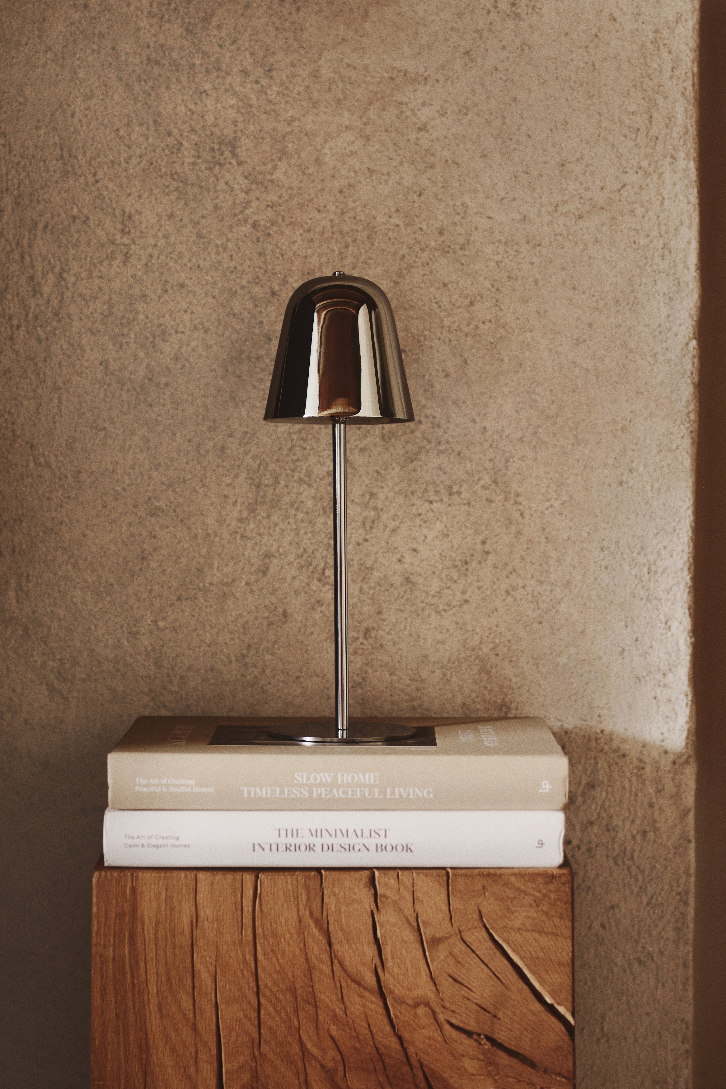 A polished chrome table lamp is positioned facing forwards, featuring a highly reflective, dome-shaped shade with a small button on top. The shade is supported by a slender, straight cylindrical stem that connects to a thin, flat circular base.