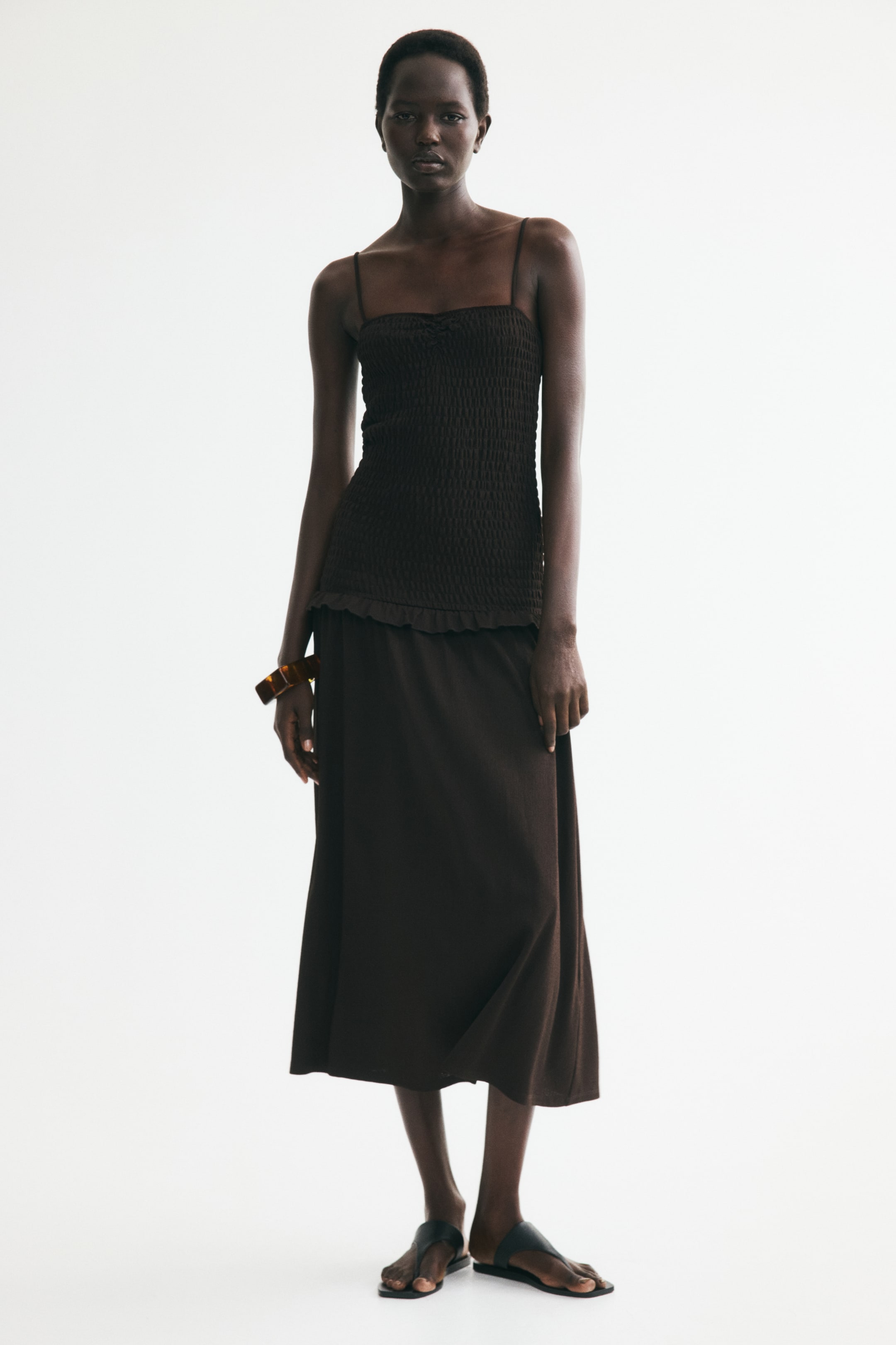 Ampliar la imagen: Un vestido de jersey a media pierna con un escote de corazón, tirantes ajustables de espagueti, un corpiño fruncido, una cintura baja adornada con volantes y una falda ligeramente acampanada está siendo lucido por la modelo.